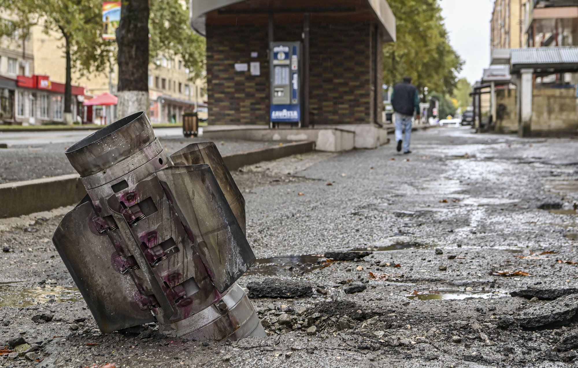 L'image montre un paysage urbain marqué par la destruction. Au premier plan, on voit un obus ou un engin explosif détérioré, partiellement enfoui dans le sol. Le bitume est endommagé autour de l'obus, laissant des fissures et des débris. En arrière-plan, un homme marche sur le trottoir, près d'un bâtiment qui semble intact. L'atmosphère est sombre et témoigne des conséquences d'un conflit ou d'un événement violent. Les arbres sur le bord de la route ajoutent une touche de verdure, mais le contexte reste très préoccupant.