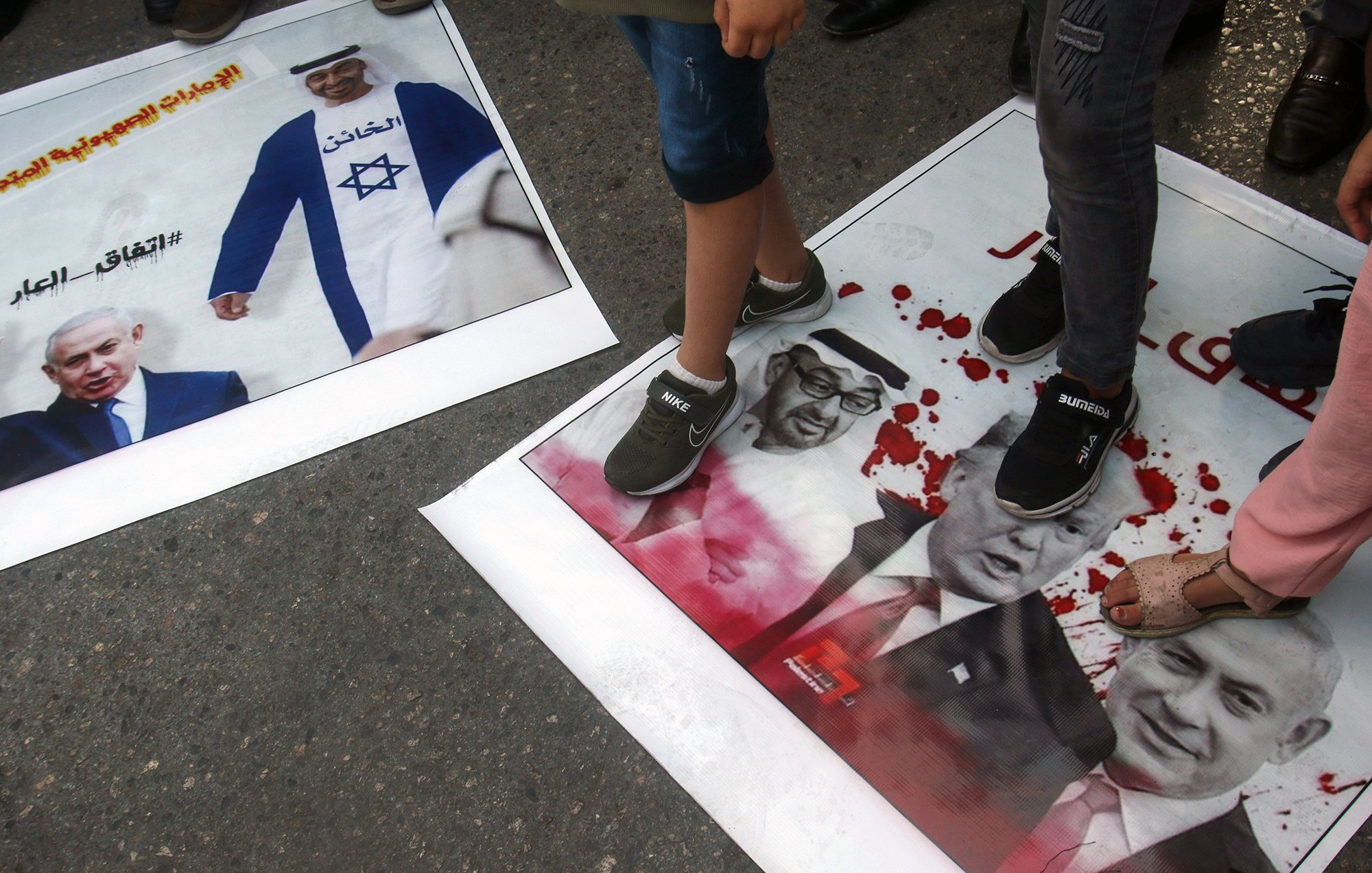 در این تصویر، تعدادی از افراد با پا بر روی پوسترهایی که عکس‌هایی از شخصیت‌های سیاسی معروف را نشان می‌دهند، ایستاده‌اند. یکی از پوسترها به تصویر نتانیاهو مربوط می‌شود و دیگری شامل تصاویری از ترامپ و یک شخصیت دیگر است. نشان‌های خون و نوشته‌ها بر روی پوسترها به وضوح دیده می‌شوند و این تصویر به نظر می‌رسد در یک تظاهرات یا اعتراض گرفته شده باشد.