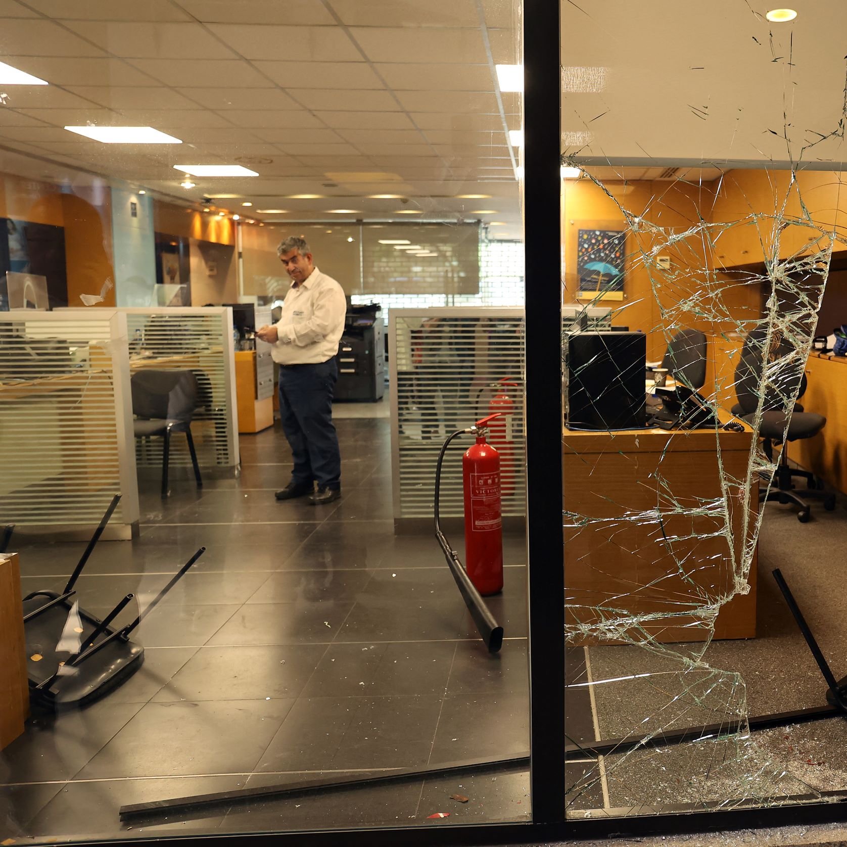 این تصویر یک نمای داخلی از یک دفترکار را نشان می‌دهد که به وضوح آسیب دیده است. شیشه‌ی پنجره شکسته و تکه‌های آن روی زمین پخش شده‌اند. در داخل اتاق چند صندلی معکوس و به هم ریخته وجود دارد. یک مرد در حال ایستادن در نزدیکی کامپیوترها به نظر می‌رسد و مشغول بررسی وضعیت است. همچنین در گوشه‌ای از اتاق، یک کپسول آتش‌نشانی قرار دارد. فضای کلی تصویر حاکی از بی‌نظمی و آسیب‌های شدید در این محیط کاری است.