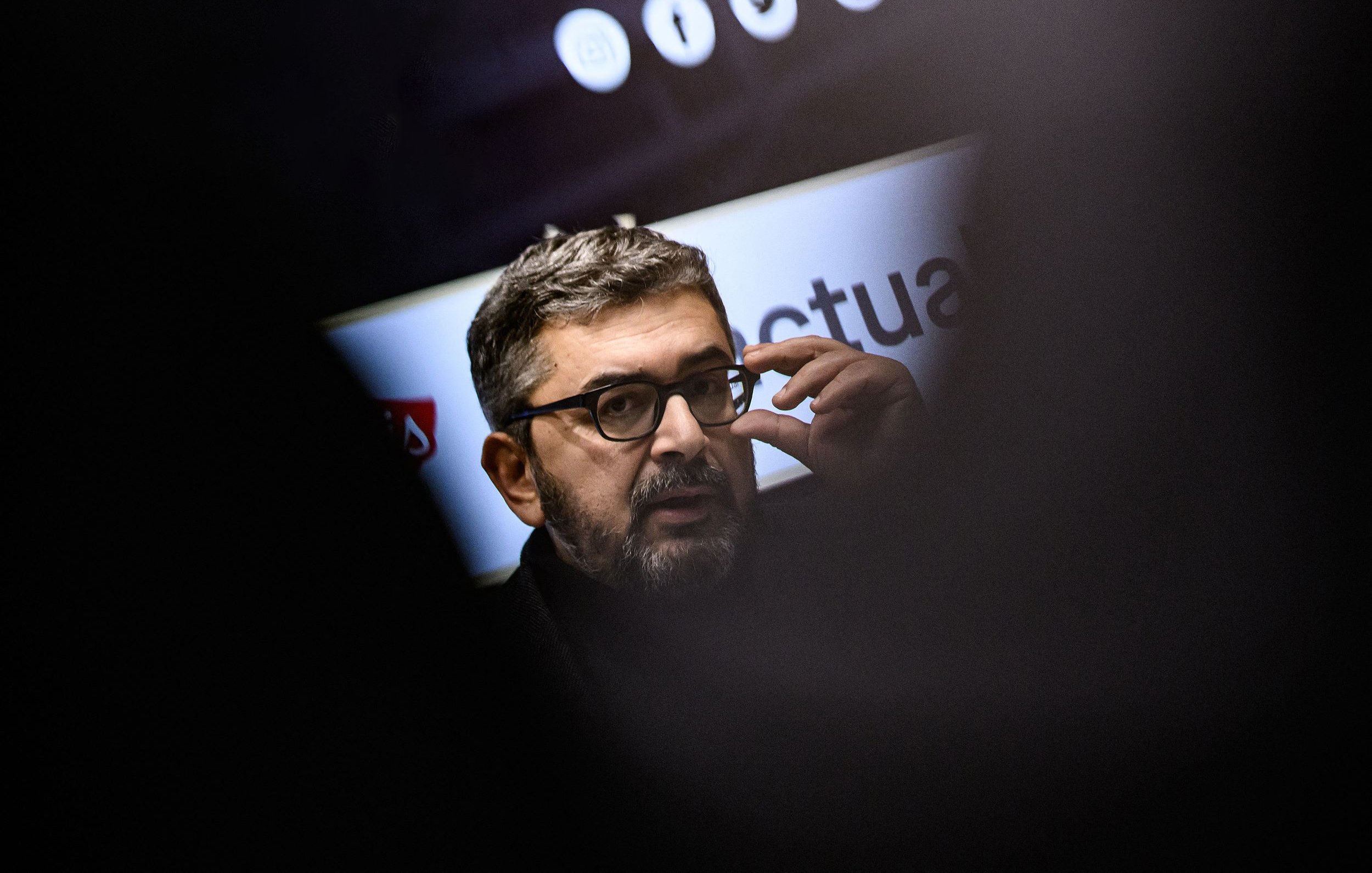 L'image montre un homme portant des lunettes et ayant une barbe courte. Il semble engagé dans une conversation ou une présentation, en touchant son visage avec une main. En arrière-plan, des éléments graphiques ou des logos sont flous, ce qui suggère qu'il se trouve dans un cadre professionnel ou public. L'éclairage met en avant son visage, tandis que des silhouettes floues d'autres personnes sont visibles autour.