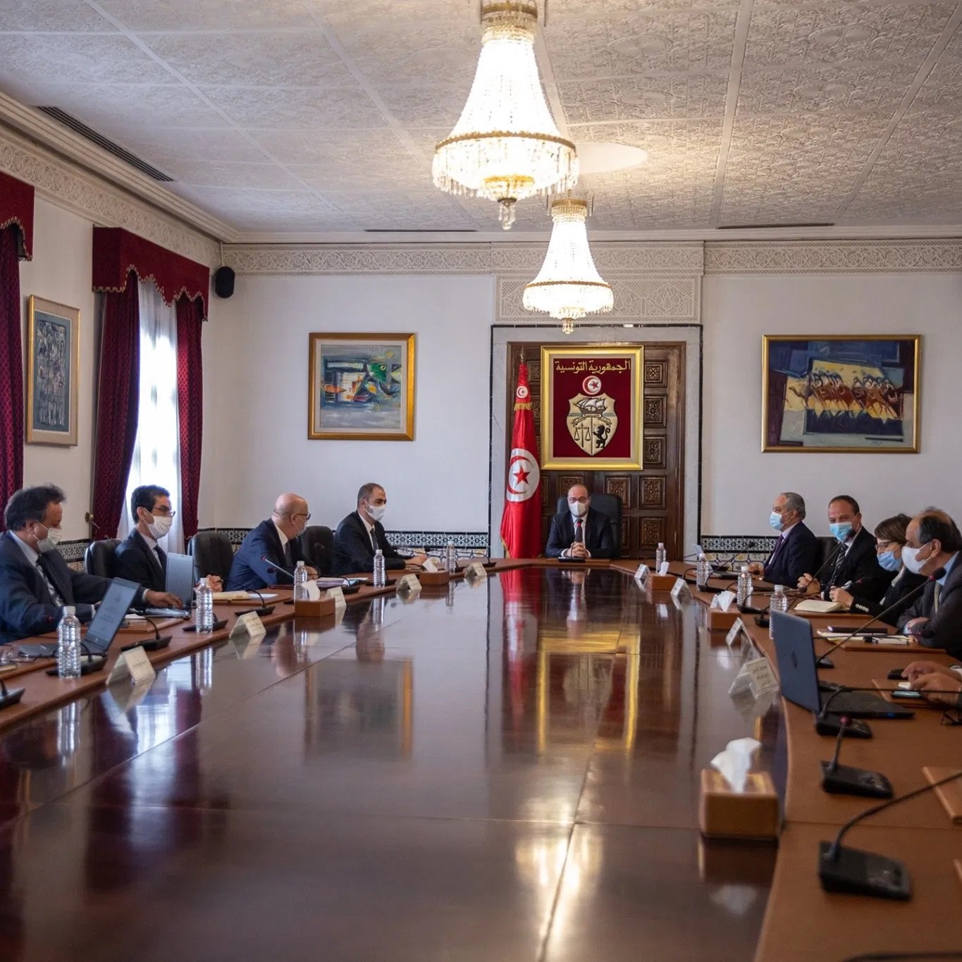 L'image montre une réunion qui se déroule dans une salle de conférence élégante. Il y a plusieurs personnes assises autour d'une grande table en bois. Au centre, un homme semble diriger la réunion, tandis que les autres participants l'écoutent attentivement. La salle est décorée de tableaux et présente des fenêtres avec des rideaux rouges. Tous les participants portent des masques, ce qui suggère une prudence sanitaire. L'atmosphère est formelle, typique d'une réunion gouvernementale ou d'affaires.