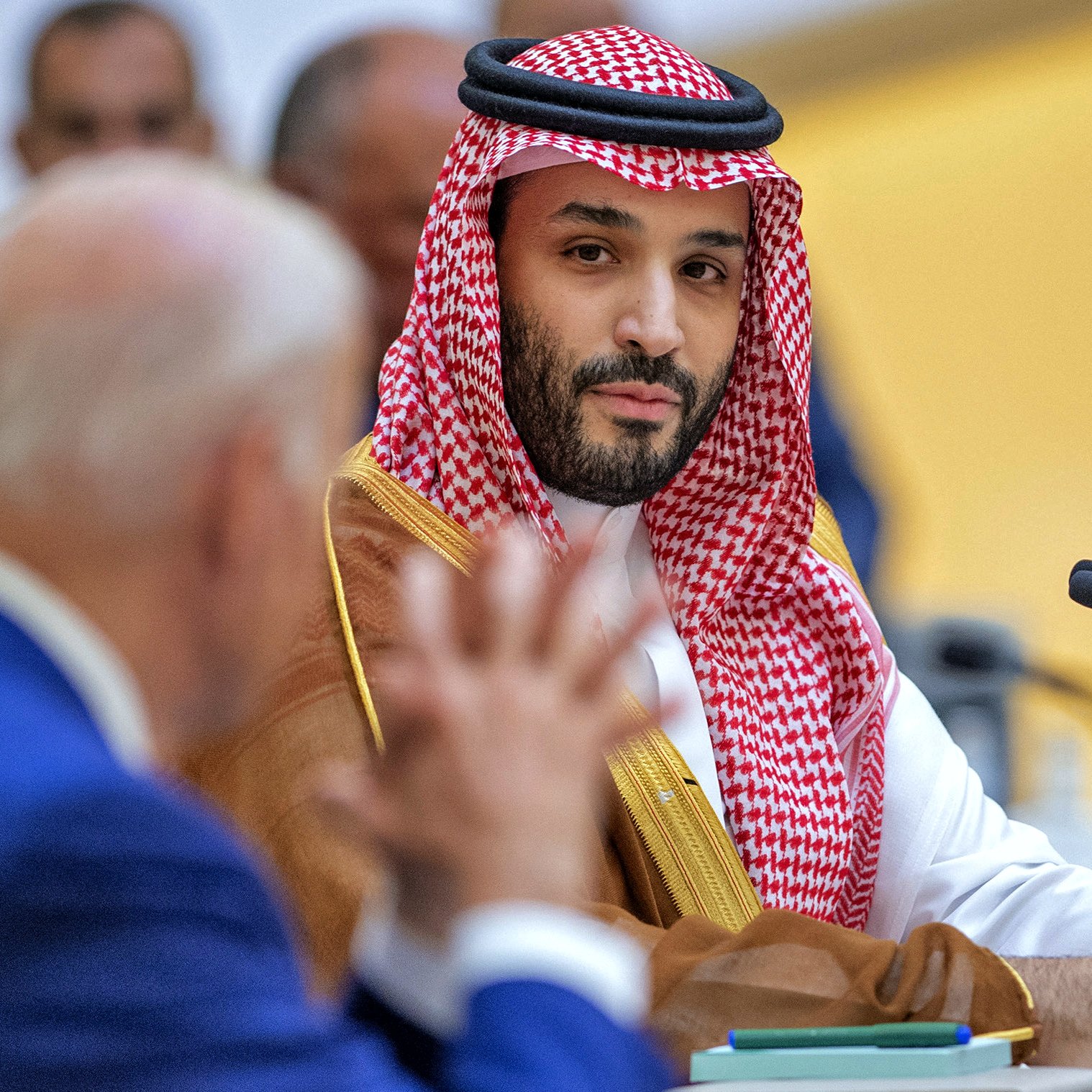 L'image montre un homme portant une tenue traditionnelle saoudienne, comprenant un thobe et un ghutrah (un couvre-chef en tissu). Il est assis à une table lors d'une réunion, fixant le spectateur avec un regard sérieux. À l'arrière-plan, un autre homme, dont le visage est partiellement visible, semble être en train de parler ou de poser une question. L'environnement suggère un cadre formel et officiel, comme une conférence ou un sommet.