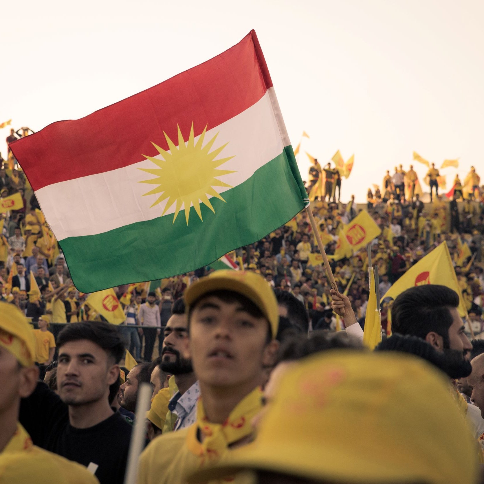 L'image montre une foule nombreuse, probablement lors d'un événement ou d'une célébration. Au premier plan, une personne brandit un drapeau kurde qui présente des couleurs vives avec un soleil stylisé. La plupart des membres de la foule portent des casquettes ou des vêtements jaunes, ajoutant à l'ambiance festive. L'arrière-plan est rempli de personnes, toutes engagées et enthousiastes, ce qui suggère une forte mobilisation et un sentiment de communauté.