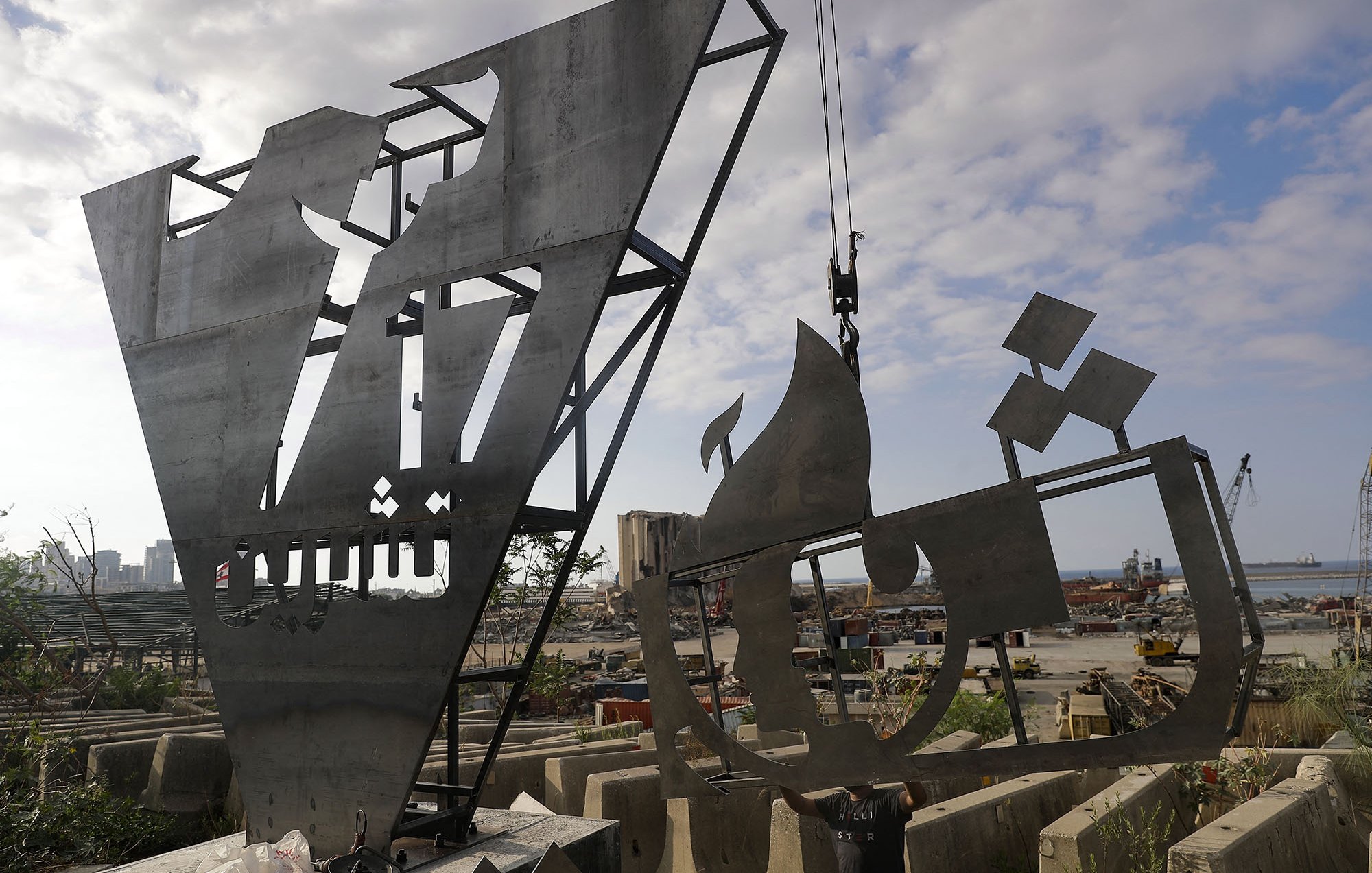 L'image montre une œuvre d'art en acier, probablement une sculpture moderne, qui est en cours d'installation ou de fabrication. La sculpture présente des formes géométriques et abstraites, avec des motifs qui semblent représenter des éléments architecturaux ou des symboles culturels. En arrière-plan, on aperçoit un chantier ou un port, avec des équipements de construction et des bateaux. Le ciel est partiellement nuageux, ajoutant une ambiance dynamique à la scène.