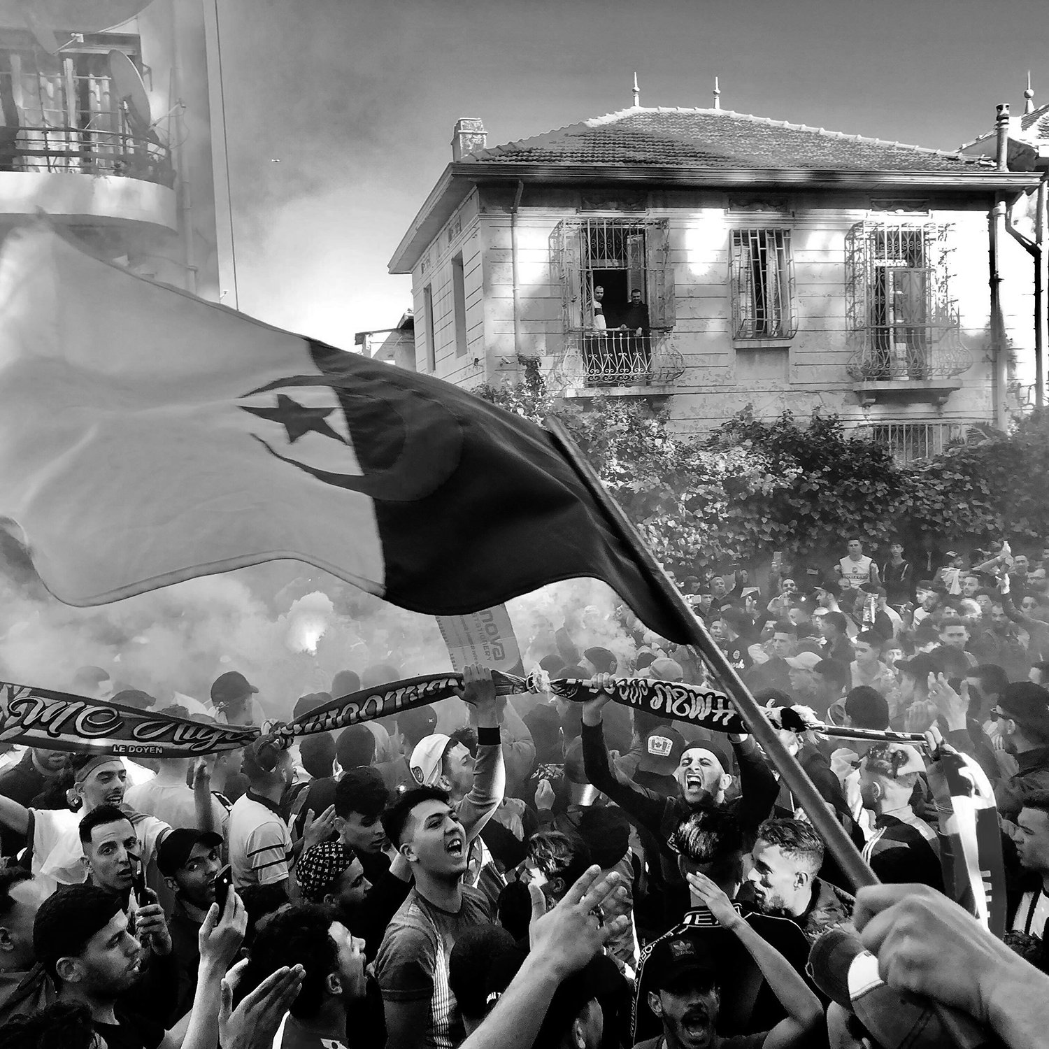 L'image montre une scène vibrante et animée, probablement lors d'un événement festif ou d'une manifestation. On peut voir une grande foule de personnes rassemblées, certaines brandissant des drapeaux et des écharpes. Les visages expriment la joie et l'enthousiasme. Une atmosphère de célébration est accentuée par des fumigènes qui créent un léger brouillard. En arrière-plan, on distingue des bâtiments, ce qui suggère que cette scène se déroule en milieu urbain.