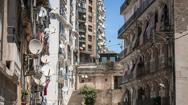 L'image montre une vue urbaine d'une rue avec des bâtiments en pierre et des murs en béton. On observe un mélange d'architecture ancienne et moderne, avec des balcons ornés de fer forgé. De nombreux paraboles satellites sont installées sur les façades des bâtiments, tandis que des linges pendus à l'extérieur ajoutent une touche de vie quotidienne. Les escaliers menant à d'autres niveaux sont visibles, et le ciel est clair et ensoleillé, donnant une atmosphère vive à la scène.