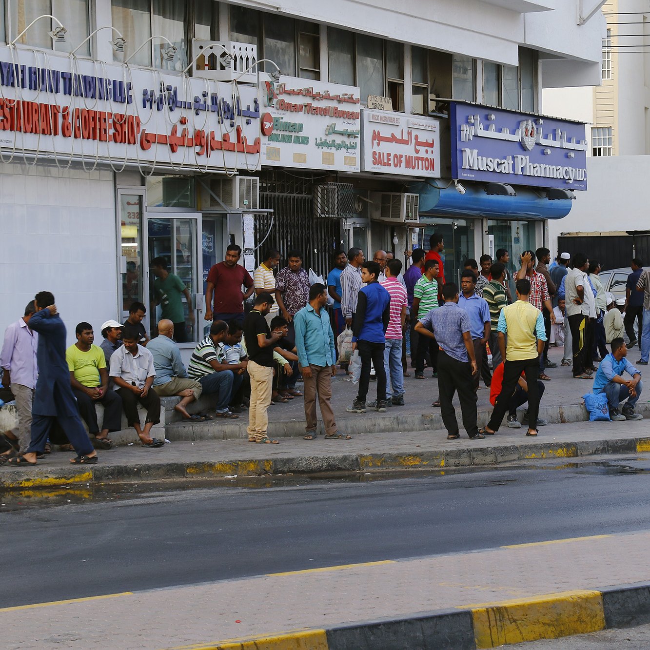 L'image montre une scène urbaine animée, avec un groupe de personnes rassemblées devant un bâtiment. On peut voir des hommes de différentes tenues discutant, assis ou se tenant debout sur le trottoir. Le bâtiment porte des enseignes de restaurants et de pharmacie, indiquant une ambiance commerçante. La route est visible au premier plan, avec une bande jaune délimitant la chaussée. L'atmosphère semble vivante, typique des zones urbaines fréquentées.