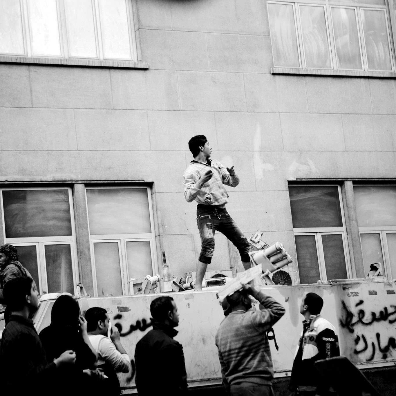 L'immagine mostra una scena di protesta in bianco e nero. Un giovane si trova in cima a un veicolo, apparentemente utilizzando un megafono o uno strumento, mentre sembra incoraggiare la folla sottostante. La gente intorno a lui è visibilmente coinvolta, alcuni alzano le mani o applaudono. Sul veicolo ci sono delle scritte, e l'architettura sullo sfondo suggerisce un contesto urbano. L'atmosfera è carica di energia e di determinazione.