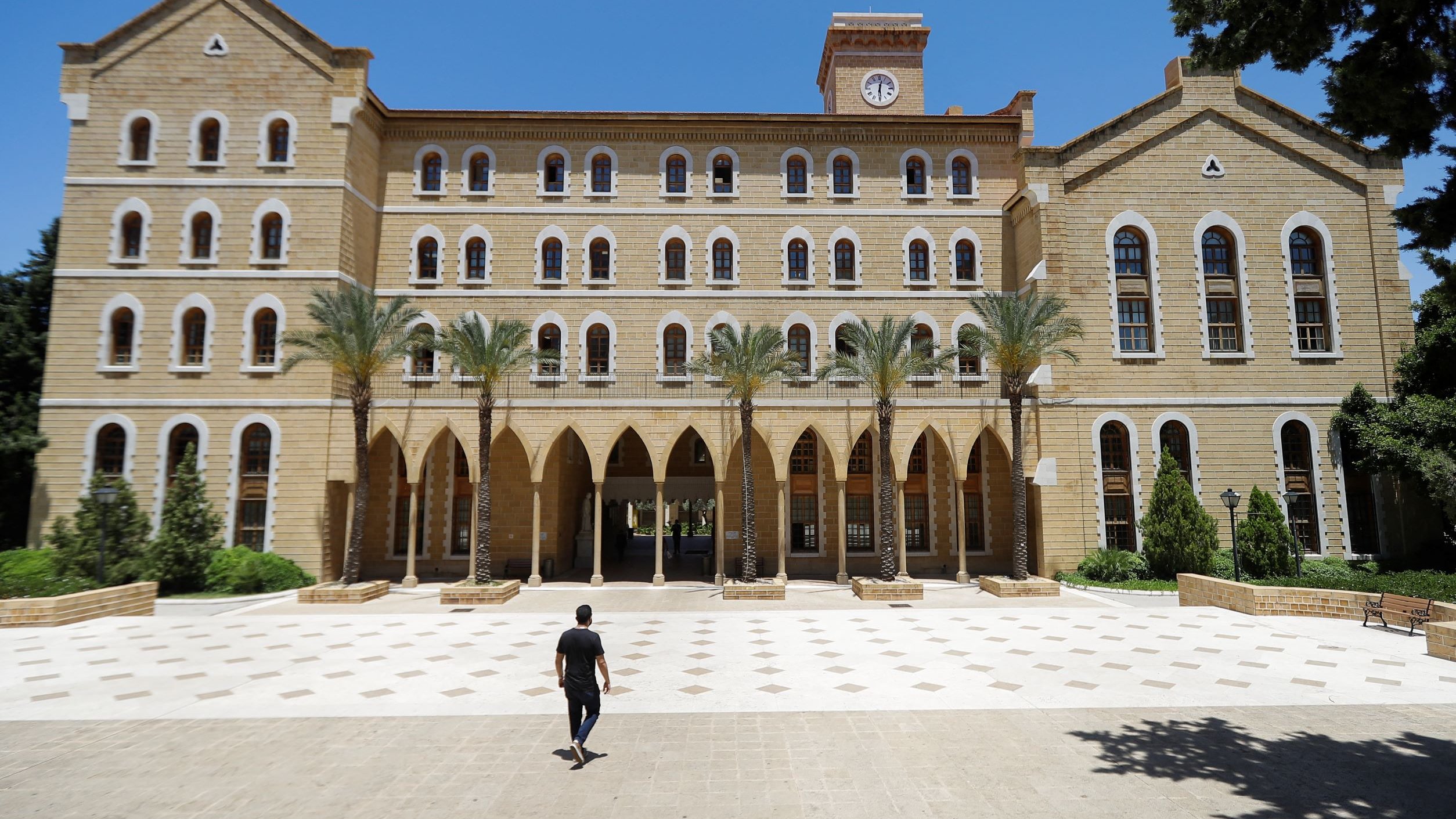 L'image montre un grand bâtiment en pierre avec une architecture traditionnelle, comportant plusieurs fenêtres à arc et des colonnes. Au premier plan, une personne marche seule sur une place pavée, tandis que des palmiers sont visibles sur les côtés. Le ciel est clair et bleu, ce qui donne une atmosphère ensoleillée. Le bâtiment semble être une institution éducative ou un lieu historique.