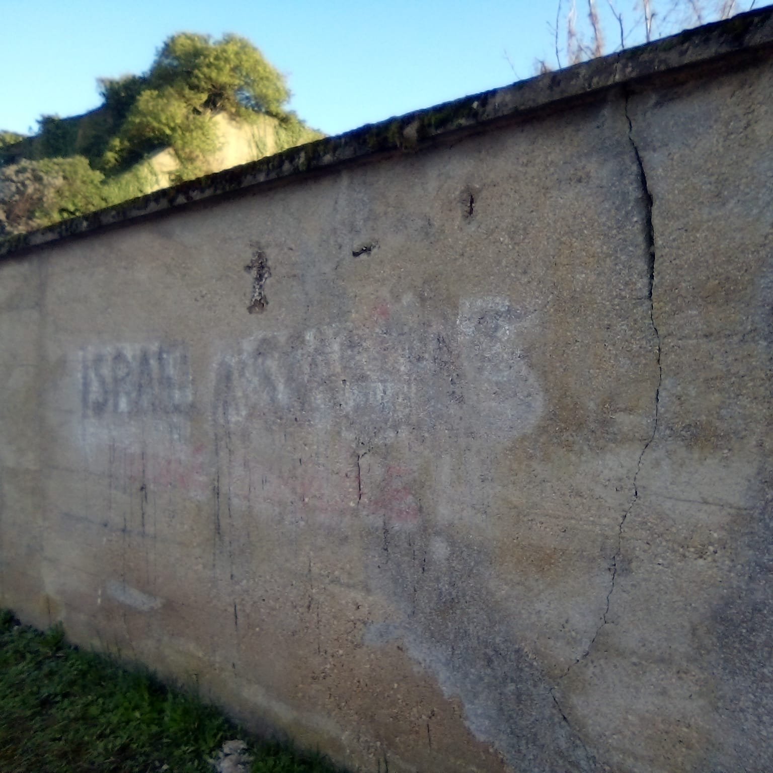 Mur en béton avec des inscriptions effacées et des fissures, sous un ciel bleu.