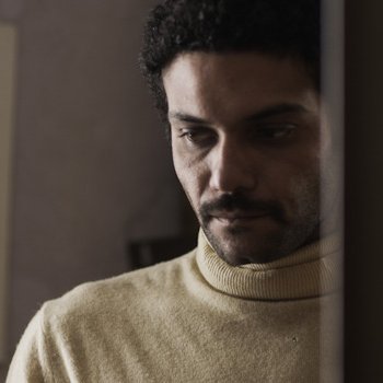 L'image montre un homme avec des cheveux bouclés et une moustache, portant un pull à col roulé de couleur claire. Il semble pensif, regardant vers le bas, avec une expression de réflexion ou de sérieux. En arrière-plan, on aperçoit des éléments de décoration intérieure, mais les détails sont flous. L'atmosphère de l'image dégage une certaine intimité ou mélancolie.