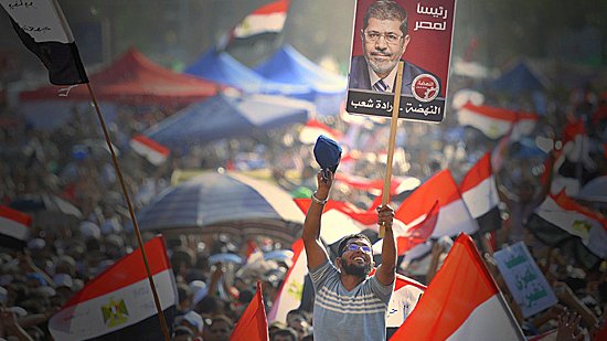 L'image montre une grande foule de manifestants brandissant des drapeaux égyptiens. Au premier plan, un homme lève une pancarte avec le portrait de Mohamed Morsi, qui était le président de l'Égypte. Les participants semblent enthousiasmés et mobilisés, reflétant une atmosphère de rassemblement populaire et d'engagement politique. Le paysage est rempli de tentes et d'une multitude de personnes, créant une impression de solidarité.