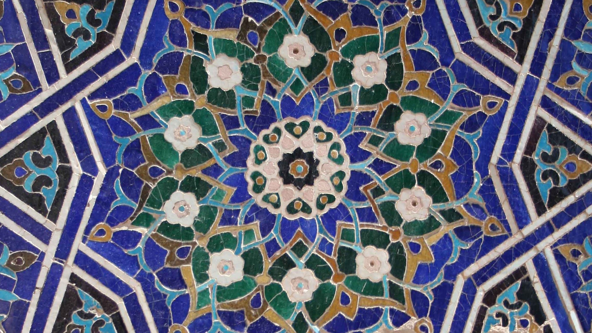 Cette image montre un motif géométrique complexe, typique de l'art islamique. On peut y voir des formes florales stylisées entourées de couleurs vives, principalement des nuances de bleu, de vert, et de blanc. Les motifs sont symétriques et créent une belle étoile centrale, ajoutant à l'aspect orné et élégant de la conception. L'ensemble véhicule une impression de délicatesse et de richesse esthétique.