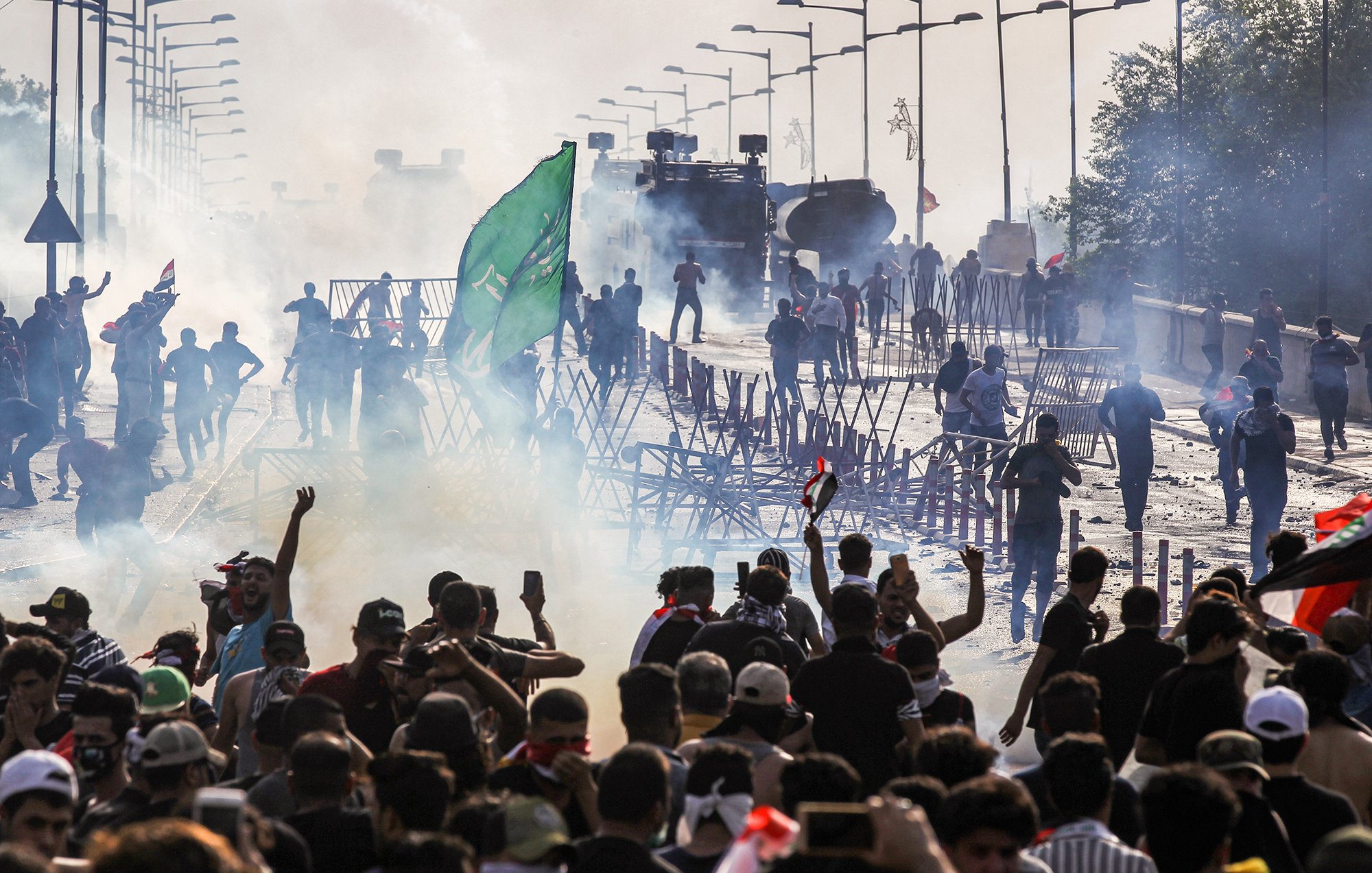 L'image montre une scène de tumultes ou de manifestations. On y voit un grand groupe de personnes rassemblées, brandissant des drapeaux et levant les bras. Il y a des barrages en bois et de la fumée, probablement causée par des gaz lacrymogènes. À l'arrière-plan, des véhicules blindés sont visibles, renforçant l'idée d'une situation tendue. L'atmosphère est chargée et chaotique, avec des manifestants qui semblent déterminés et en mouvement.
