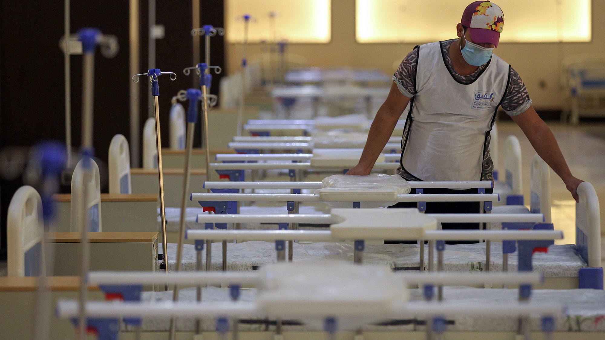 L'image montre une personne qui se trouve dans un environnement médical, probablement un hôpital ou un centre de soins. Cette personne porte un masque et un gilet, et elle est en train de s'occuper de plusieurs lits médicaux alignés. Les lits sont équipés de draps et de dispositifs intraveineux. L'atmosphère semble calme et organisée, suggérant un espace dédié aux soins des patients.