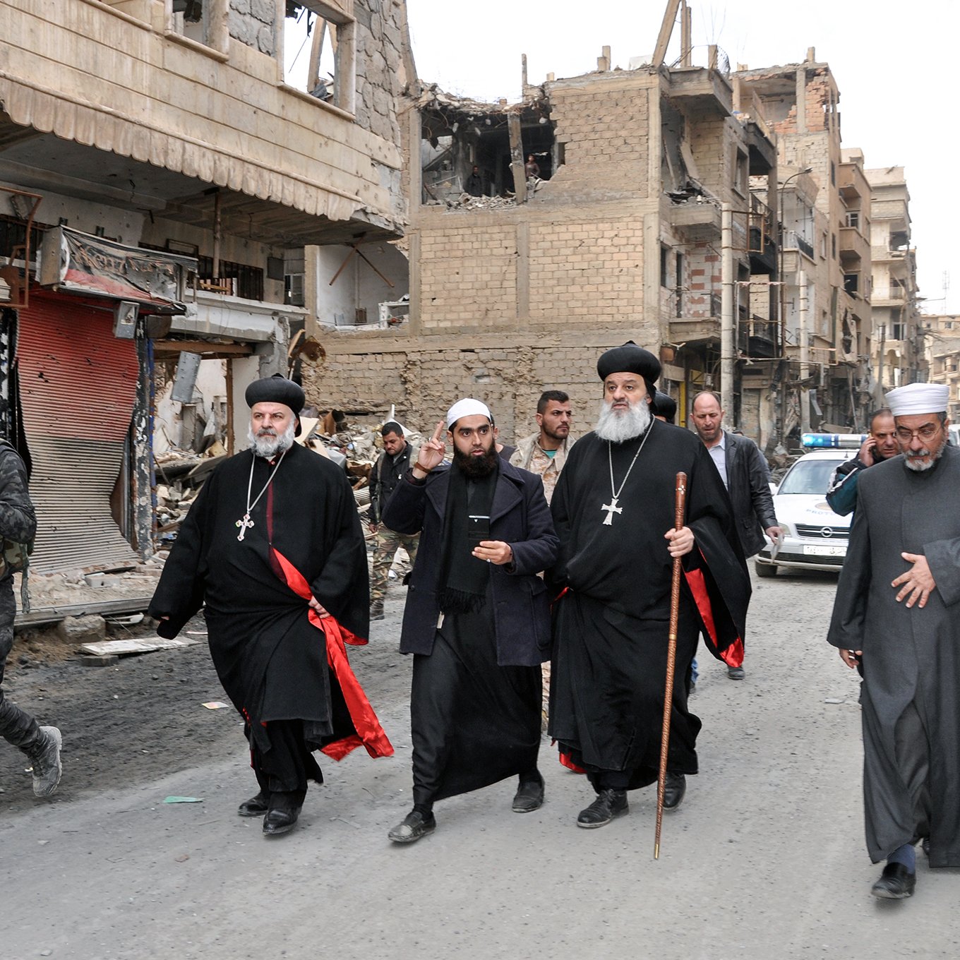 L'image montre un groupe de personnes marchant dans une rue endommagée. Il y a des bâtiments en ruines sur les côtés, indiquant une zone touchée par des conflits. Parmi le groupe, plusieurs hommes portent des vêtements traditionnels, notamment des robes noires et des croix, ce qui suggère qu'ils pourraient être des leaders religieux. Certains d'entre eux tiennent des bâtons. En arrière-plan, des militaires et des véhicules sont visibles, ajoutant à l'ambiance de la scène.