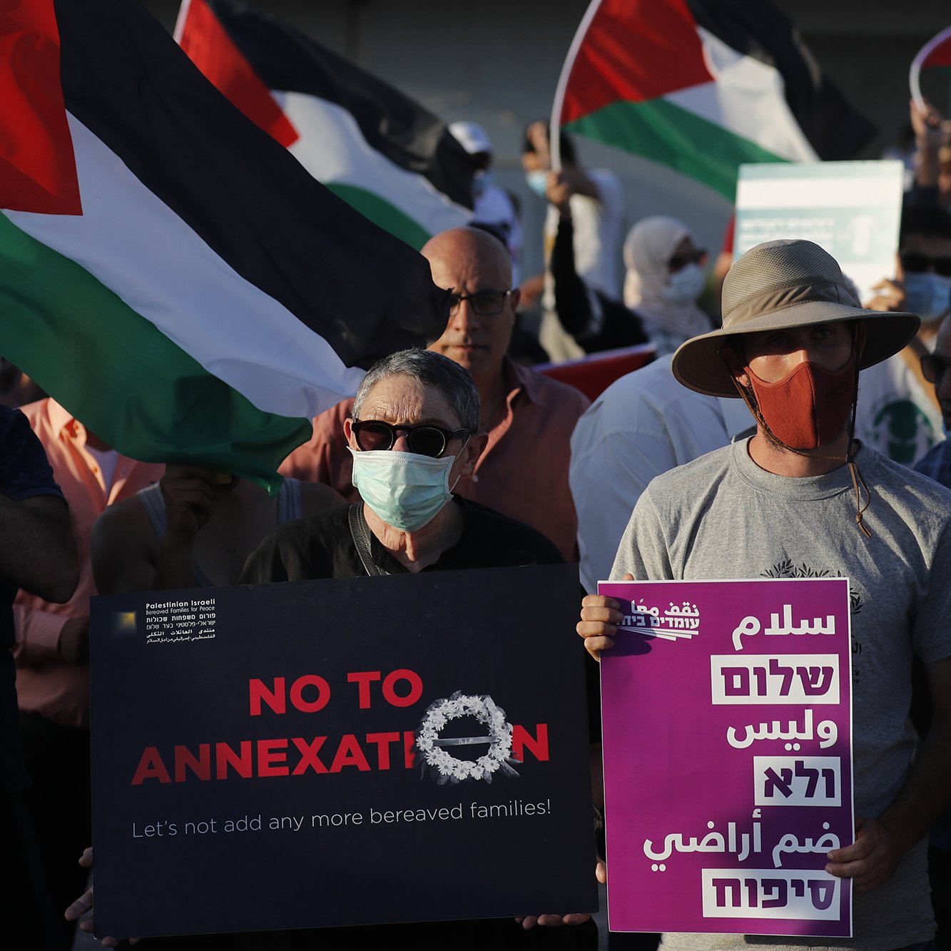 در این تصویر، گروهی از مردم در حال برگزاری یک تظاهرات هستند. آنها پرچم‌های فلسطینی را در دست دارند و تعدادی از آنها تابلوهای اعتراضی حمل می‌کنند. نوشته‌های روی تابلوها شامل عباراتی مانند "نه به الحاق" و "بیایید خانواده‌های بیشتری را اضافه نکنیم" به چشم می‌خورد. همچنین، برخی از تظاهرکنندگان ماسک به صورت دارند. فضای تصویر به وضوح نشان‌دهنده اعتراض و حمایت از حقوق فلسطینیان است.