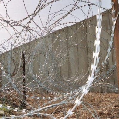 L'image montre un paysage marqué par des barbelés disposés en spirale et un mur en béton en arrière-plan. Le terrain est rocheux et dépouillé, ce qui contribue à une ambiance austère et sécurisée. Les barbelés créent une impression de barricade ou de clôture, suggérant une séparation ou un isolement. Le ciel au-dessus est clair, ajoutant un contraste avec les éléments de sécurité présents dans le paysage.