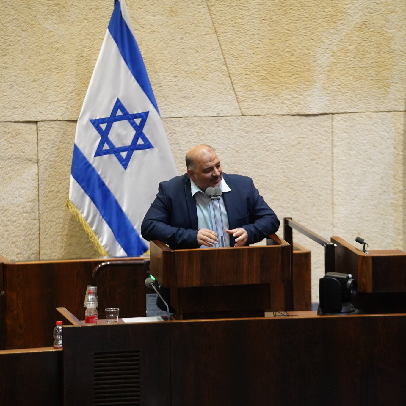 L'image montre un homme debout à un pupitre dans une salle de réunion, probablement au sein d'une assemblée ou d'un parlement, avec des drapeaux israéliens en arrière-plan. Le décor est sobre, avec des murs en pierre et du mobilier en bois. L'homme semble s'adresser à un public, exprimant peut-être son opinion sur un sujet d'actualité.
