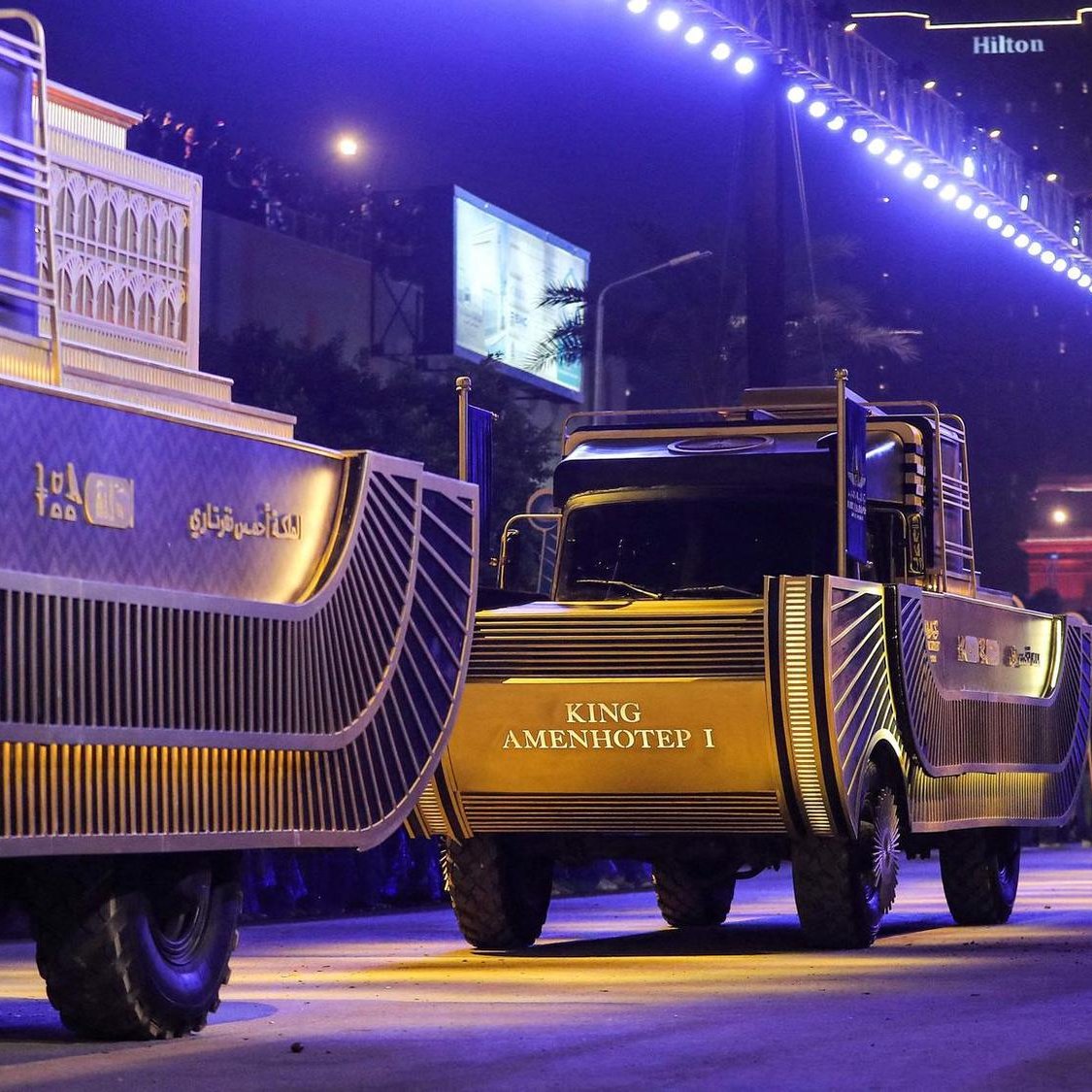 L'image montre un défilé nocturne avec des camions décorés, probablement pour une célébration ou un événement spécial. Les camions portent des inscriptions, dont "KING AMENHOTEP I", et sont éclairés par des lumières vives. En arrière-plan, on aperçoit un bâtiment, peut-être un hôtel, et des foules observant l'événement. Un personnage en costume traditionnel est également visible, ajoutant à l'atmosphère festive et culturelle.