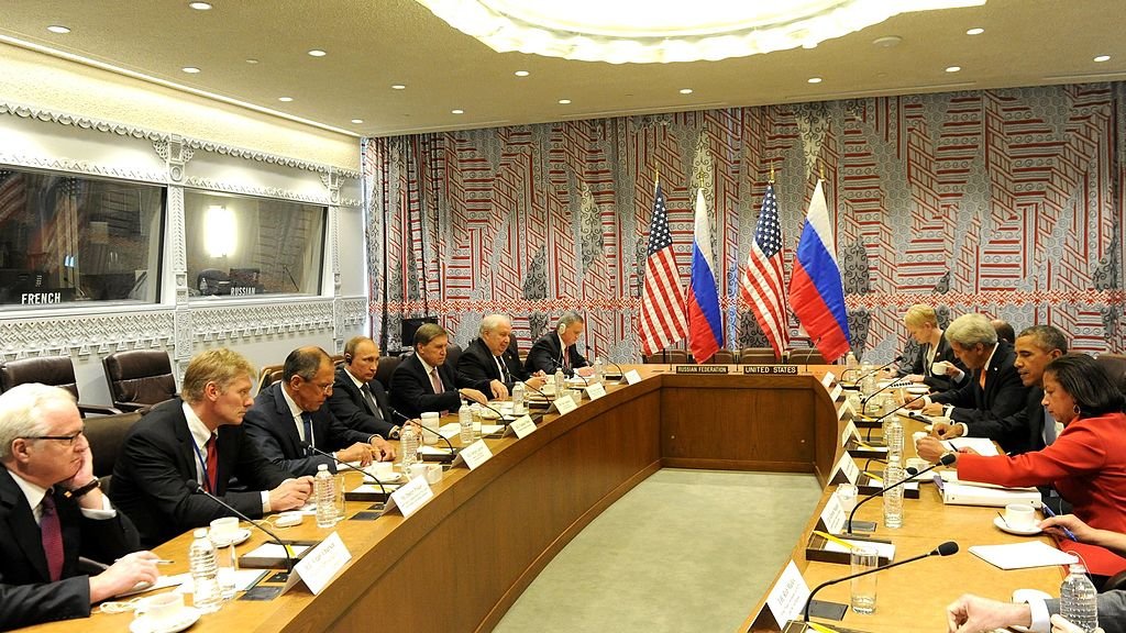 تظهر في الصورة مجموعة من المسؤولين في قاعة اجتماعات. الجانبان يمثلان الولايات المتحدة وروسيا، مع وجود علمي كلا البلدين على الطاولة. يبدو أن الاجتماع يتناول قضايا دبلوماسية أو سياسية هامة. الطاولة طويلة، وهناك عدد من الكراسي المحيطة بها، حيث يجلس المشاركون يتبادلون النقاشات. تظهر ملامح الجدية والتركيز على وجوههم، مما يدل على أهمية الحوار الجاري. الديكور الخلفي للمكان يتضمن تصاميم حديثة.
