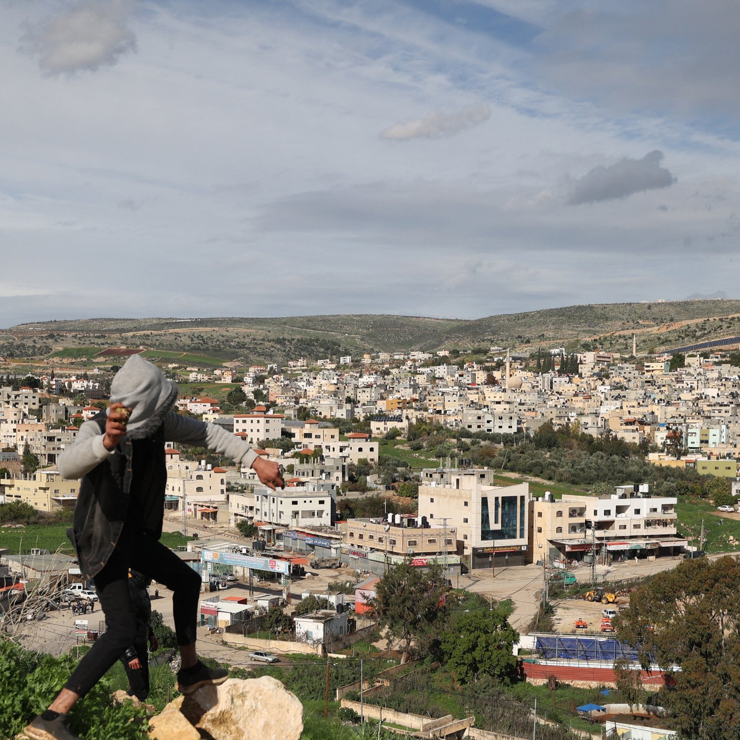L'image montre une vue panoramique d'un paysage urbain avec de nombreux bâtiments construits, probablement dans une région montagneuse ou vallonnée. Au premier plan, une personne est en train de sauter ou de se déplacer sur des rochers. L'horizon est marqué par des collines et des nuages, et on peut voir des routes et des structures en dessous, indiquant une zone habitée. La scène dégage une atmosphère dynamique, avec un mélange d'éléments naturels et urbains.