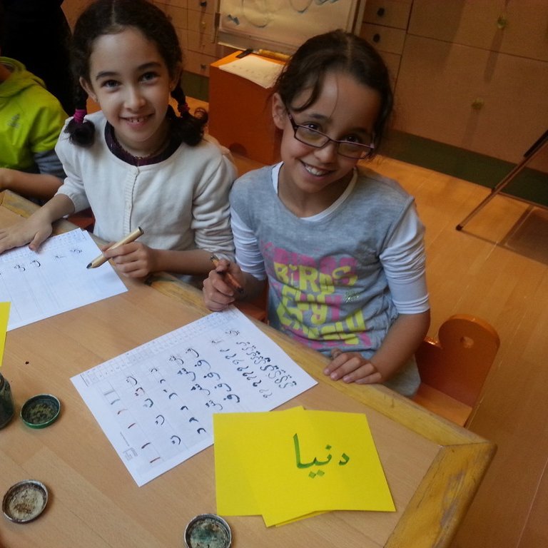 L'image montre deux jeunes filles assises à une table, souriant et travaillant sur des feuilles de papier. Elles utilisent des pinceaux et de l'encre, et semblent engagées dans une activité créative, probablement de calligraphie. Des étiquettes jaunes sont visibles, sur l'une est écrit "سيماء" et sur l'autre "دنيا" en arabe. L'environnement est scolaire, avec d'autres enfants visibles en arrière-plan.