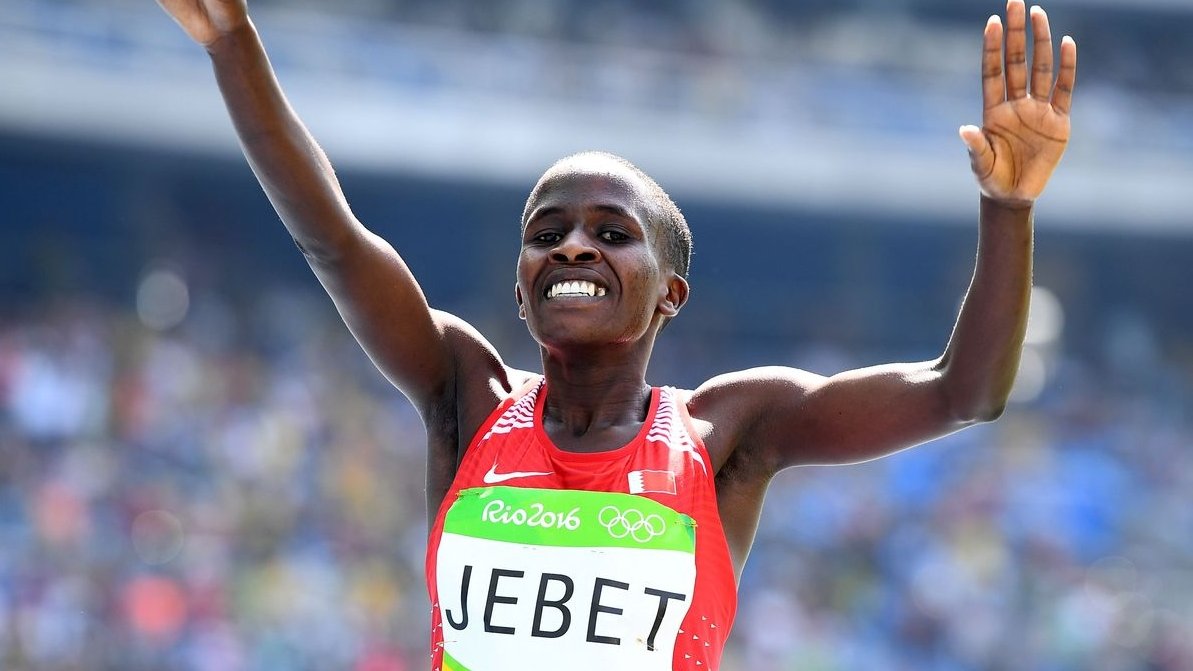 L'image montre une athlète en plein moment de célébration. Elle a les bras levés en l'air, affichant un large sourire, ce qui suggère qu'elle vient de réaliser une performance exceptionnelle. Elle porte un maillot rouge avec son nom "JEBET" inscrit dessus, ainsi que des éléments des Jeux Olympiques de Rio 2016. L'arrière-plan montre une foule et un stade, indiquant qu'elle est en compétition dans un événement majeur.