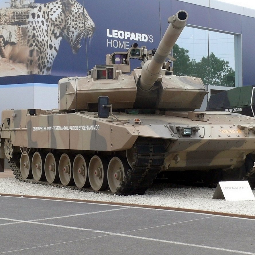 L'image montre un véhicule militaire, plus précisément un char de combat. Le char est peint dans un motif de camouflage beige et brun, typique pour se fondre dans des environnements désertiques. Il est équipé d'un canon long monté sur le dessus et possède plusieurs roues de chaque côté. En arrière-plan, on aperçoit des éléments d'exposition militaire, suggérant que le char est présenté lors d'un salon ou d'une exposition lié à la défense. Les détails tels que les plaques d'identification du char peuvent également être visibles.