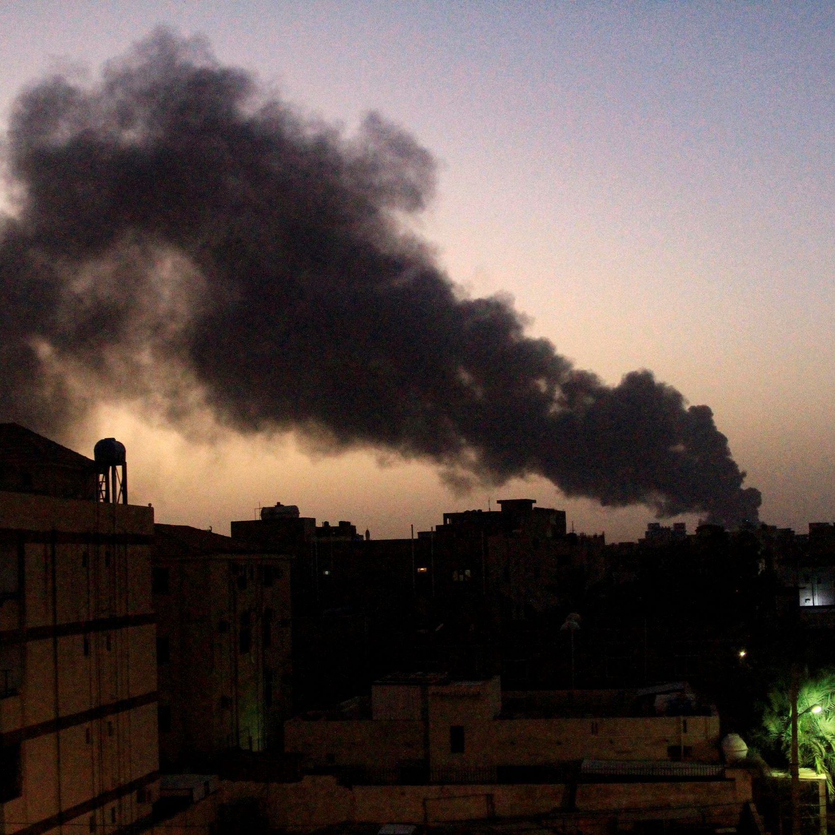 L'image montre un paysage urbain de nuit, avec des bâtiments au premier plan. En arrière-plan, de grands nuages de fumée noire s'élèvent dans le ciel, créant un contraste avec l'horizon teinté de lumière douce. On peut observer une ambiance sombre et impressionnante, suggérant une situation potentiellement problématique ou alarmante. Les silhouettes des bâtiments et les arbres ajoutent de la profondeur à la scène.