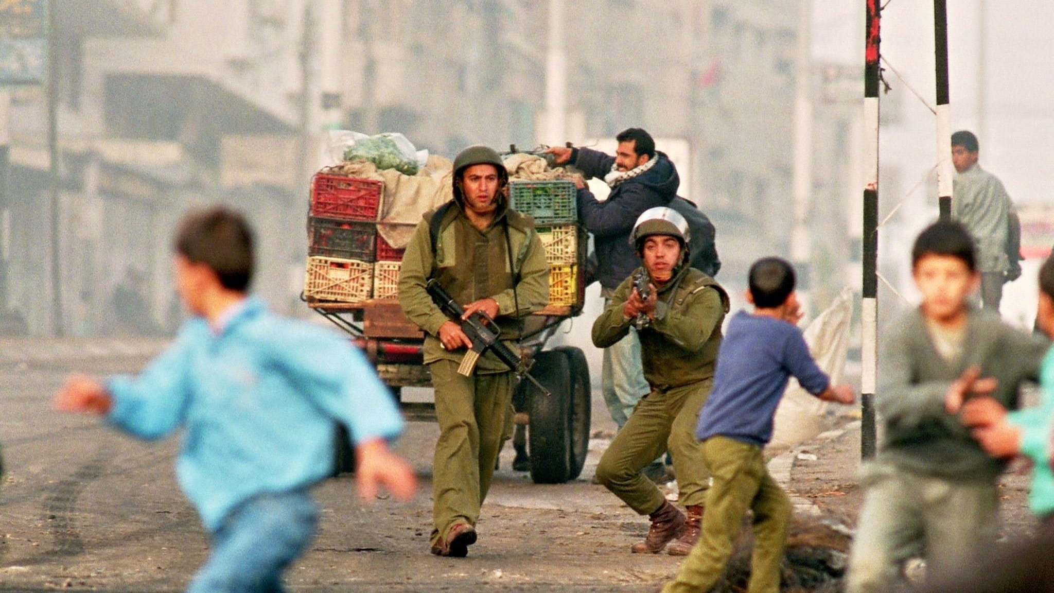L'immagine ritrae una scena di conflitto in un'area urbana. Due soldati in uniforme stanno correndo, uno dei quali sembra puntare un'arma. Dietro di loro, un carro con casse di frutta è visibile. In primo piano, dei bambini stanno fuggendo, mostrando un'espressione di paura. L'ambiente circostante appare caotico, con una luce che suggerisce un'atmosfera di tensione. La scena comunica chiaramente una situazione di crisi e instabilità.