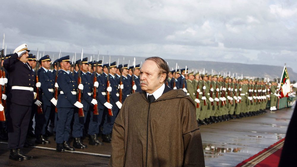 L'image montre un homme en manteau se tenant devant un alignement de soldats en uniforme. Les soldats, majestueux et bien rangés, saluent. L'arrière-plan est nuageux, créant une atmosphère cérémonielle. Le sol semble humide, indiquant peut-être qu'il a plu récemment. L'homme au premier plan a une expression pensive, ajoutant un ton sérieux à la scène.