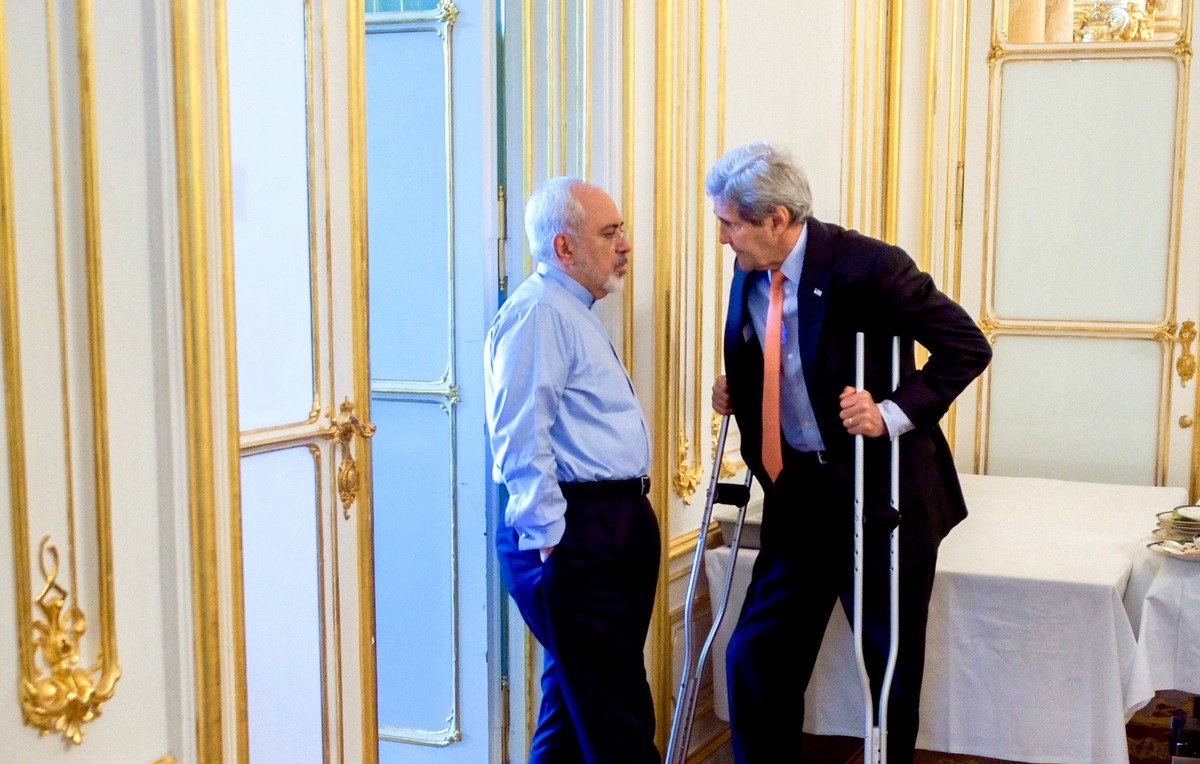 L'image montre deux hommes en train de discuter dans un environnement intérieur. L'un des hommes porte une chemise bleue et semble écouter attentivement, tandis que l'autre, utilisant des béquilles, parle avec animation. L'arrière-plan est décoré avec des moulures dorées et il y a une table couverte d'une nappe blanche. L'atmosphère semble informative et sérieuse.