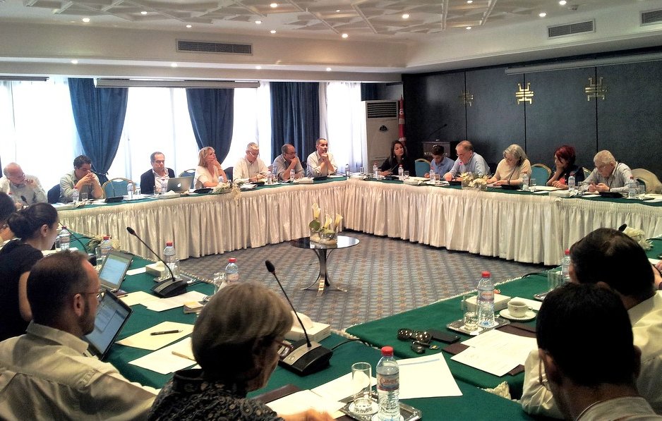 L'image montre une grande salle de réunion où se déroule une conférence ou un séminaire. Plusieurs personnes sont assises autour d'une table ovale, certaines utilisant des ordinateurs portables. Les participants semblent engagés dans des discussions, et il y a des documents et des bouteilles d'eau sur chaque table. La pièce est bien éclairée par la lumière naturelle qui entre par de grandes fenêtres, et des rideaux bleus ajoutent une touche décorative. Le décor est professionnel, avec des éléments modernes et une ambiance de collaboration.