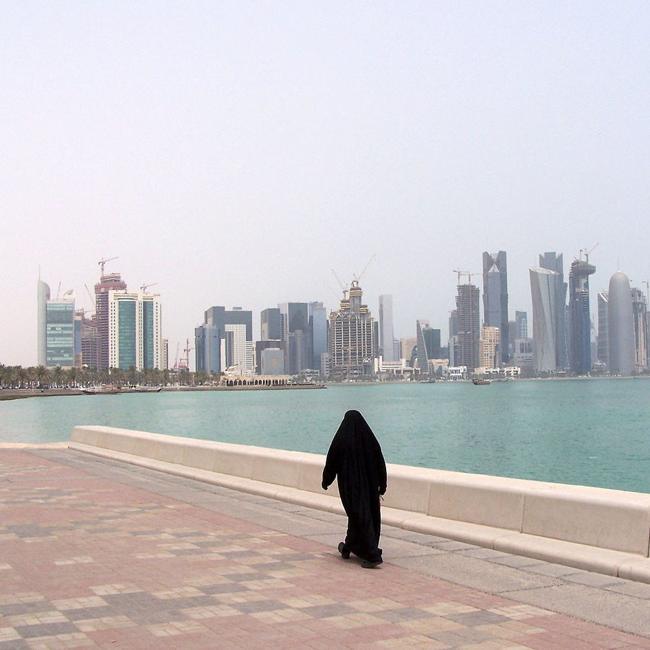 L'image montre une personne vêtue d'une tenue traditionnelle noire, marchant le long d'une promenade au bord de l'eau. En arrière-plan, on aperçoit une ligne d'horizon urbaine avec des gratte-ciel modernes et des bâtiments en construction, représentant une ville côtière. L'ambiance est calme, avec un ciel légèrement brumeux et une mer tranquille. Les pavés de la promenade ajoutent une touche de couleur à la scène.