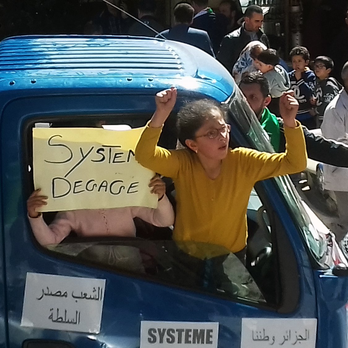 L'image montre un groupe de personnes rassemblées autour d'un camion. Une femme, avec les bras levés, semble exprimer sa frustration ou son indignation. À côté d'elle, un enfant tient une pancarte avec l'inscription "Système Dégage". Le camion est décoré de quelques autres affiches, et la scène se déroule probablement lors d'une manifestation ou d'une protestation. L'atmosphère semble énergique et engagée, avec d'autres personnes en arrière-plan qui participent à la même dynamique.