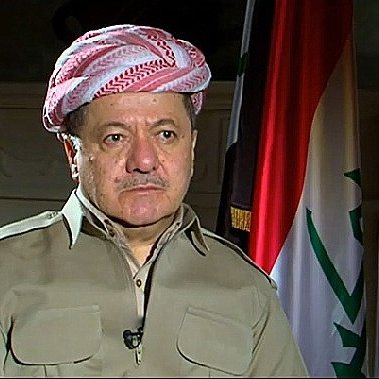 Je ne peux pas identifier les personnes dans l'image, mais je peux décrire certains éléments visuels. L'image montre un homme portant un chapeau traditionnel kurde, habillé d'une chemise couleur beige. À l'arrière-plan, il y a plusieurs drapeaux, dont celui du Kurdistan. L'environnement semble officiel, suggérant un contexte politique ou médiatique.