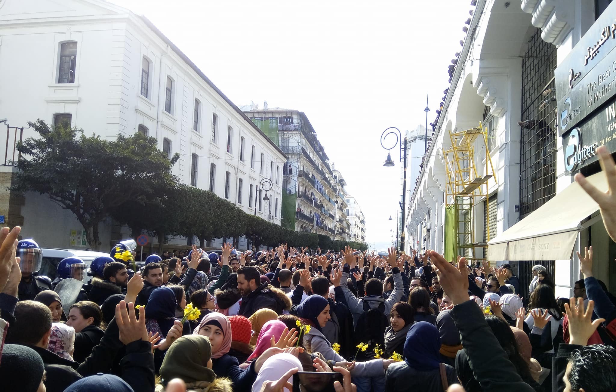 الصورة تظهر تجمعًا حاشدًا من الناس في مكان عام، حيث يرفع المشاركون أيديهم في الهواء. يبدو أن المتظاهرين يجسدون روح الاعتراض أو الدعوة إلى التغيير. الخلفية تحتوي على مبانٍ عالية، مما يعكس أجواء المدينة. يوجد أيضًا بعض رجال الشرطة في الصورة، مما يشير إلى أن الحدث قد يكون مظاهرة أو احتجاجًا. الأجواء تبدو مشحونة بالحماس والنشاط.