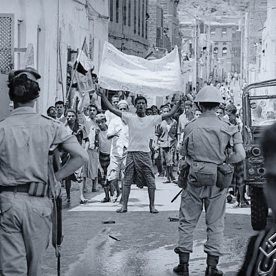 L'image montre une scène de rue dans un contexte de manifestation ou de rassemblement. On peut voir des groupes de personnes, certains brandissant des drapeaux ou des pancartes, exprimant visiblement leur émotion ou leur revendication. Des policiers réglementent la situation, et l'atmosphère semble tendue. L'environnement urbain est composé de bâtiments simples, typiques d'une ville. Les vêtements des gens indiquent peut-être une époque passée, soulignant le contexte historique de la scène.