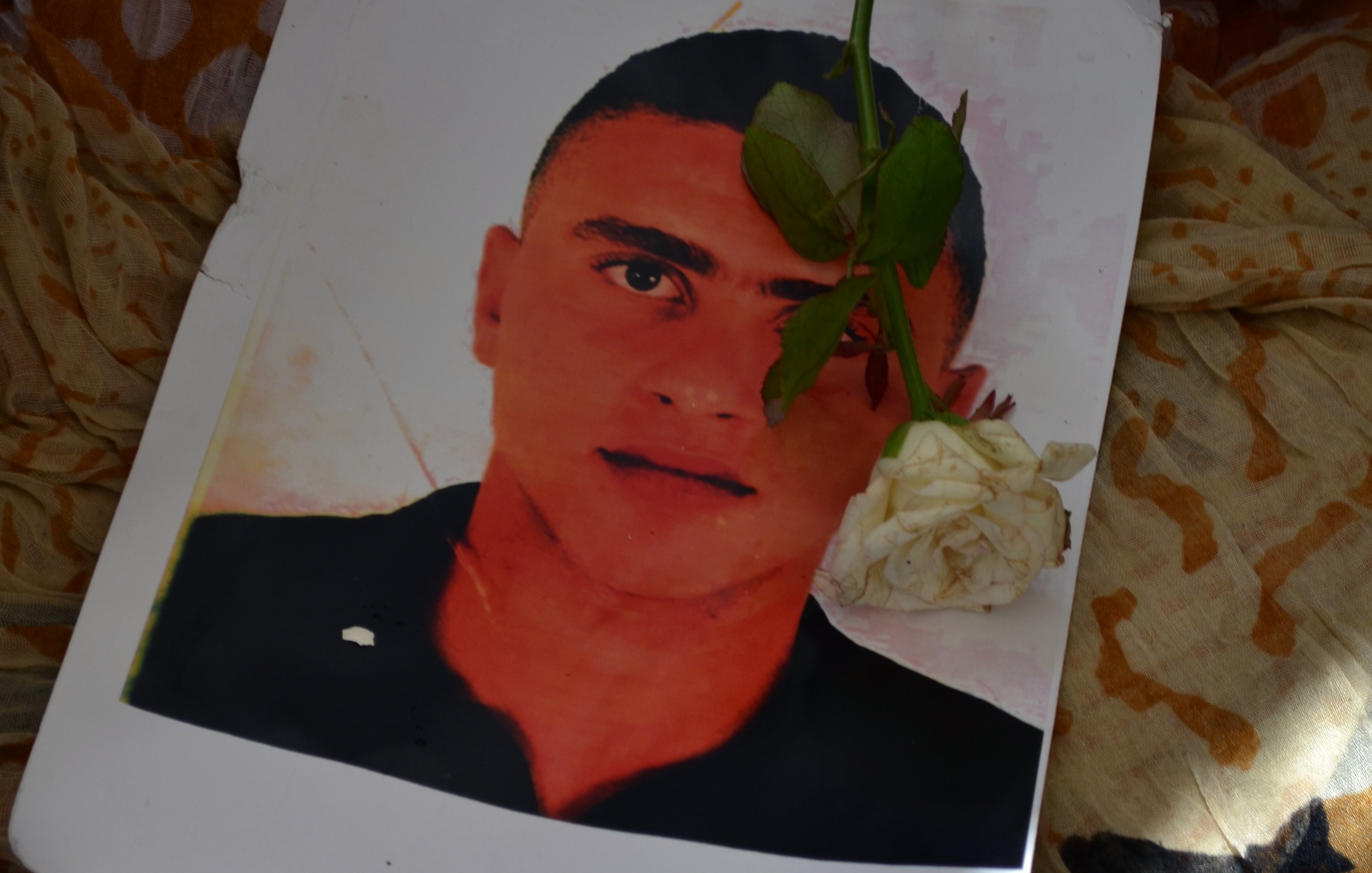 L'image montre un portrait d'un jeune homme, placé sur un tissu aux motifs colorés. Une rose blanche est délicatement posée sur le portrait, ajoutant une touche de sérénité et de respect. Les couleurs et les textures du tissu environnant contrastent avec l'image du jeune homme.