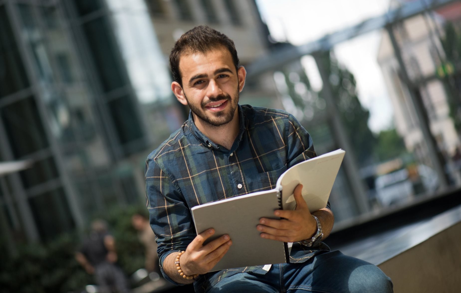 L'image montre un jeune homme assis sur un banc, souriant, tout en tenant un carnet ou un cahier dans ses mains. Il porte une chemise à carreaux et a l'air détendu, avec un environnement urbain en arrière-plan et des bâtiments modernes. La lumière naturelle éclaire la scène, créant une atmosphère agréable.
