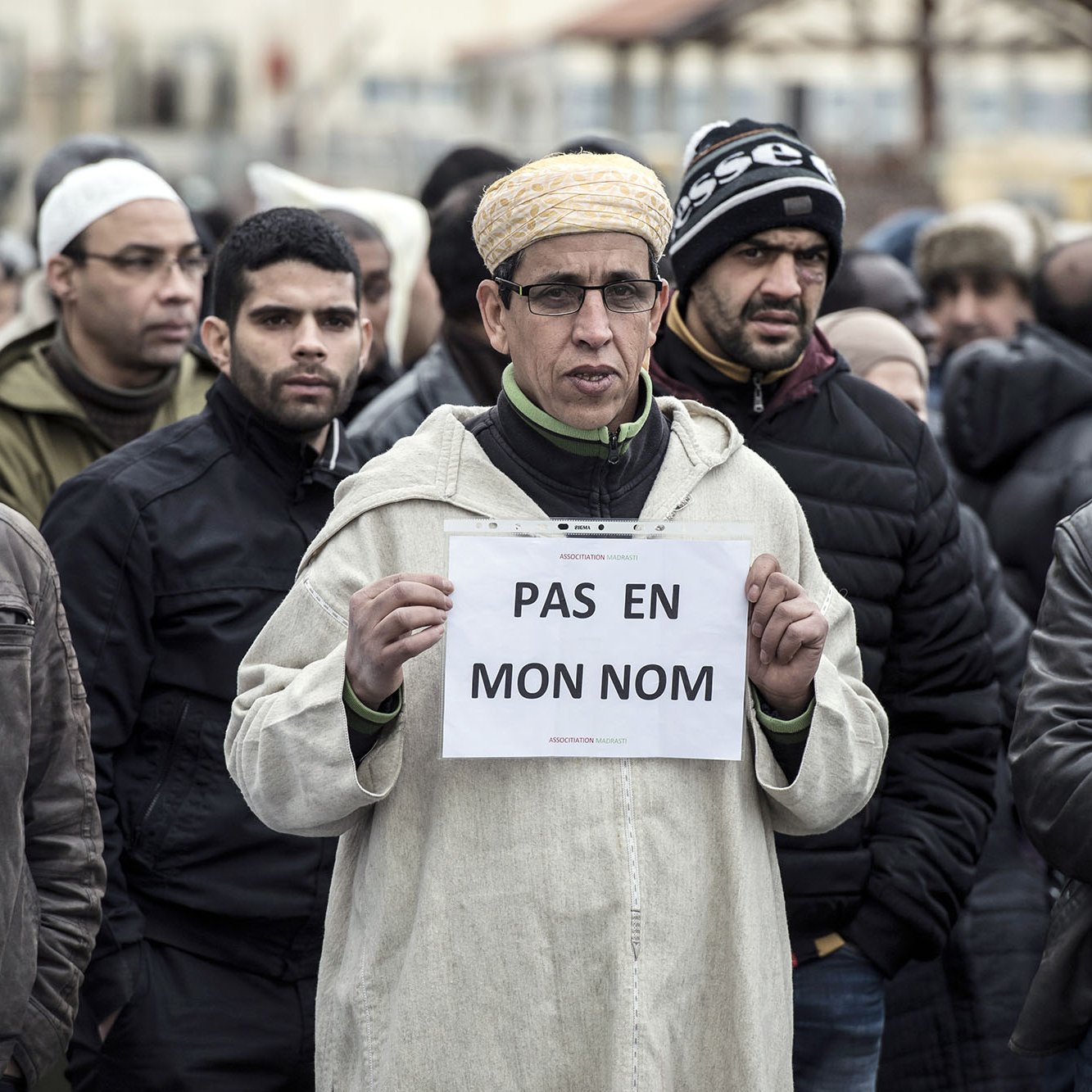 L'image montre un groupe de personnes rassemblées, dont certaines portent des vêtements d'hiver. Au premier plan, un homme se distingue en tenant une pancarte sur laquelle est écrit « PAS EN MON NOM ». Son expression est sérieuse et déterminée. Les autres personnes autour de lui semblent également concentrées sur la situation. L'ambiance générale de la scène suggère une manifestation ou un rassemblement pour une cause particulière.