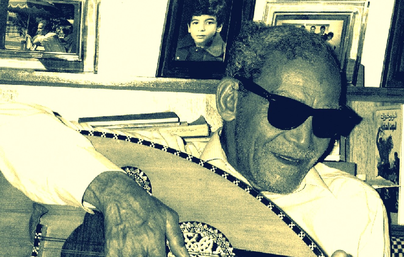 الصورة تظهر رجلًا مسنًا يرتدي نظارات شمسية ويعزف على آلة العود. يبدو سعيدًا ومسترخيًا، محاطًا بإطارات صور على الجدار خلفه. الأسلوب الذي يقدّم به العزف يعكس شغفه بالموسيقى. الأجواء تدل على لمسة من الح nostalgia، مع تفاصيل تعكس العناية والحنين إلى الماضي.