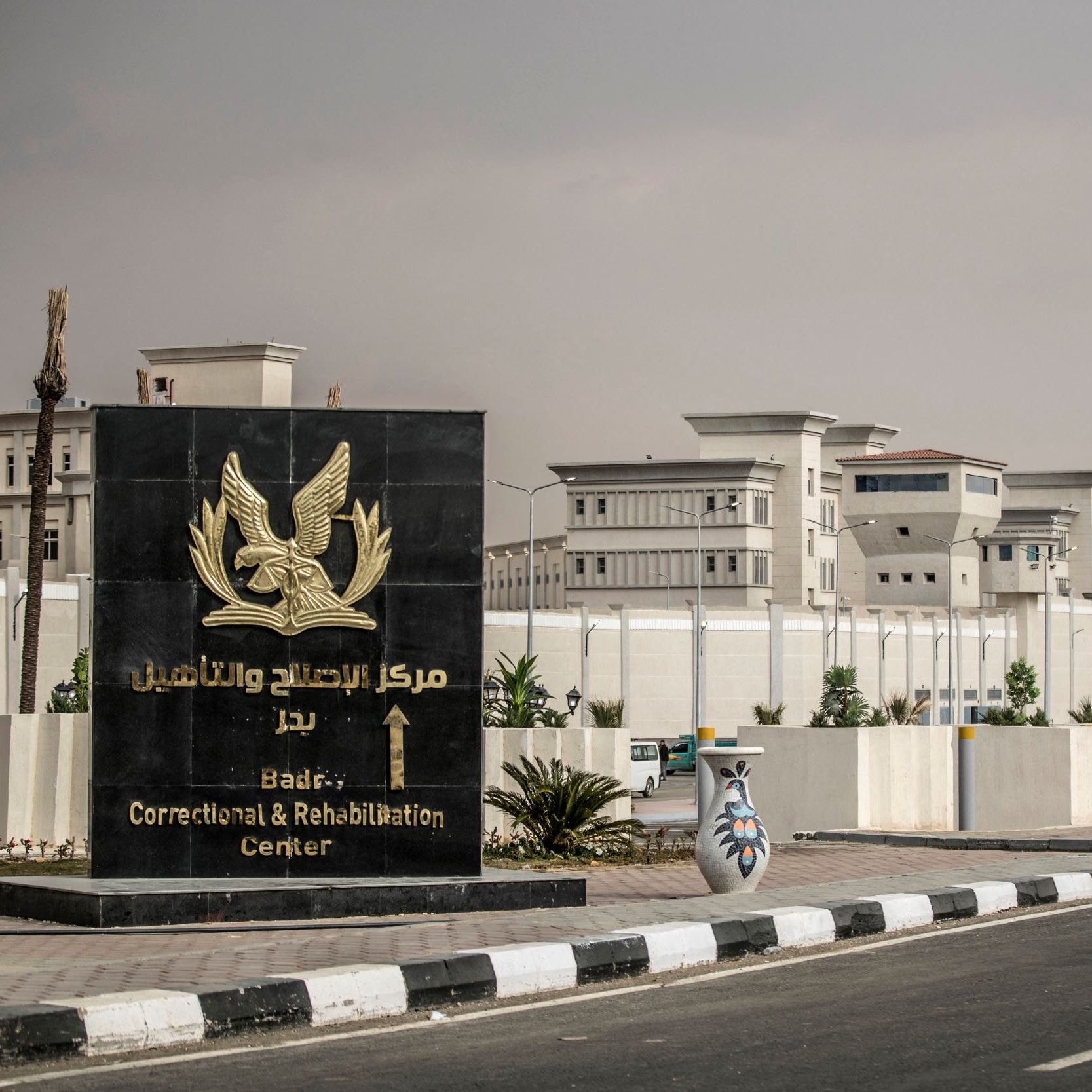 The image depicts a large sign featuring an eagle emblem, which is located in front of a correctional and rehabilitation center. The sign is written in both Arabic and English, indicating the name of the facility, which is "Badry Correctional and Rehabilitation Center." The center itself is visible in the background, showcasing a well-maintained structure surrounded by a tall wall and clear skies. There are decorative elements at the base of the sign, including ornamental vases, and the area is lined with street lamps.