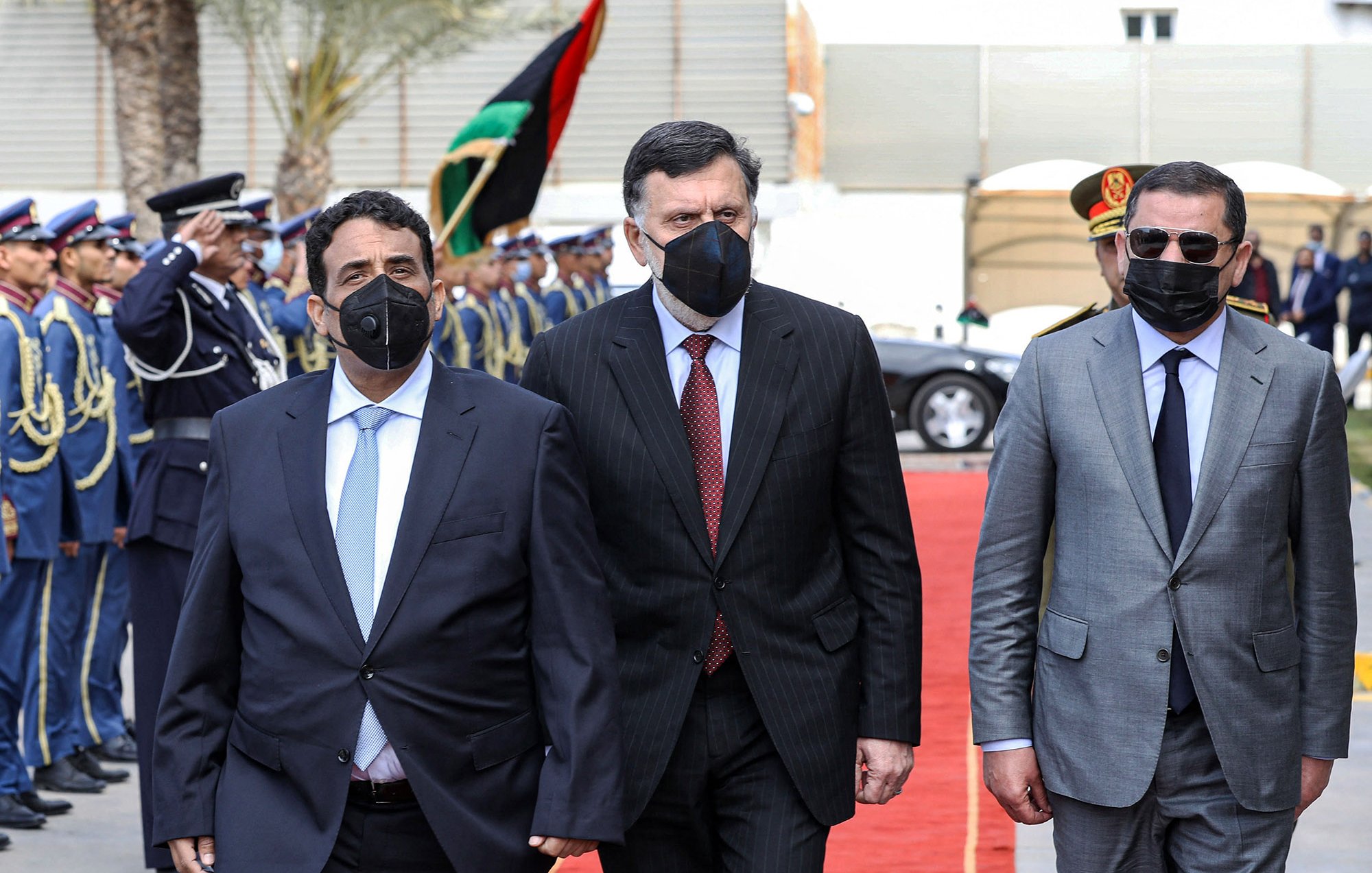 L'image montre un groupe de trois hommes marchant sur un tapis rouge, apparemment lors d'un événement officiel. Ils portent tous des costumes, et deux d'entre eux portent des masques faciaux noirs. En arrière-plan, on peut voir une garde d'honneur en uniforme, ainsi que des drapeaux. L'atmosphère semble solennelle et accueillante, suggérant une réception ou une cérémonie diplomatique.
