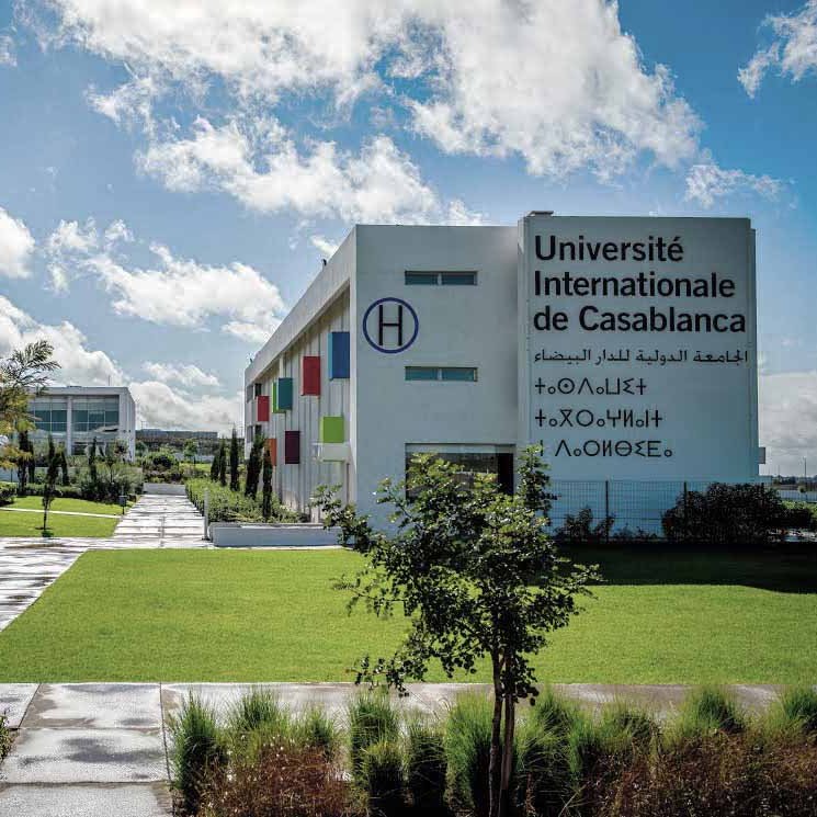 L'image montre l'Université Internationale de Casablanca. On peut voir des bâtiments modernes, avec des façades blanches et colorées. L'environnement est entouré de verdure, incluant des arbres et des espaces verts bien entretenus. Le ciel est bleu avec quelques nuages, et le sol semble humide, suggérant qu'il a peut-être récemment plu. L'ensemble dég dég dég dég dég dég dég dég dég dég dég dég dég dég dég dég dég dég dég dég dég dég dég dég dég dég deg d'une atmosphère accueillante et dynamique.