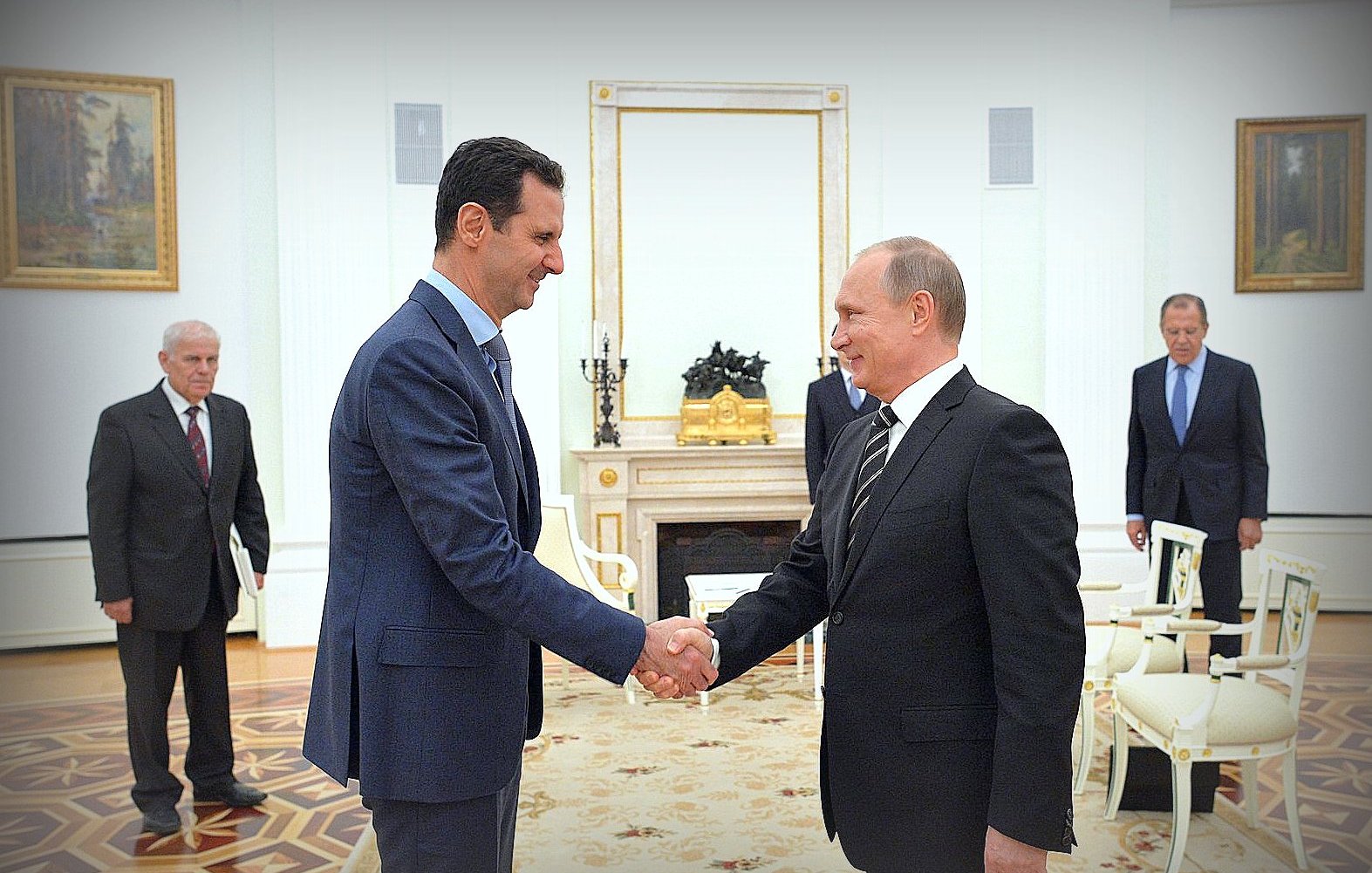 الصورة تظهر لقاءً بين شخصين رئيسيين، حيث يقوم أحدهما بمصافحة الآخر. الشخص على اليسار يرتدي بدلة زرقاء، بينما الشخص على اليمين يرتدي بدلة داكنة. يبدو أن اللقاء يجري في مكان رسمي، مع ديكور فخم بالإضافة إلى لوحات فنية على الجدران. في الخلفية، يوجد عدد من الأشخاص الآخرين الذين يشاهدون اللقاء بشكل غير مباشر. الجو العام يوحي بأهمية المناسبة.