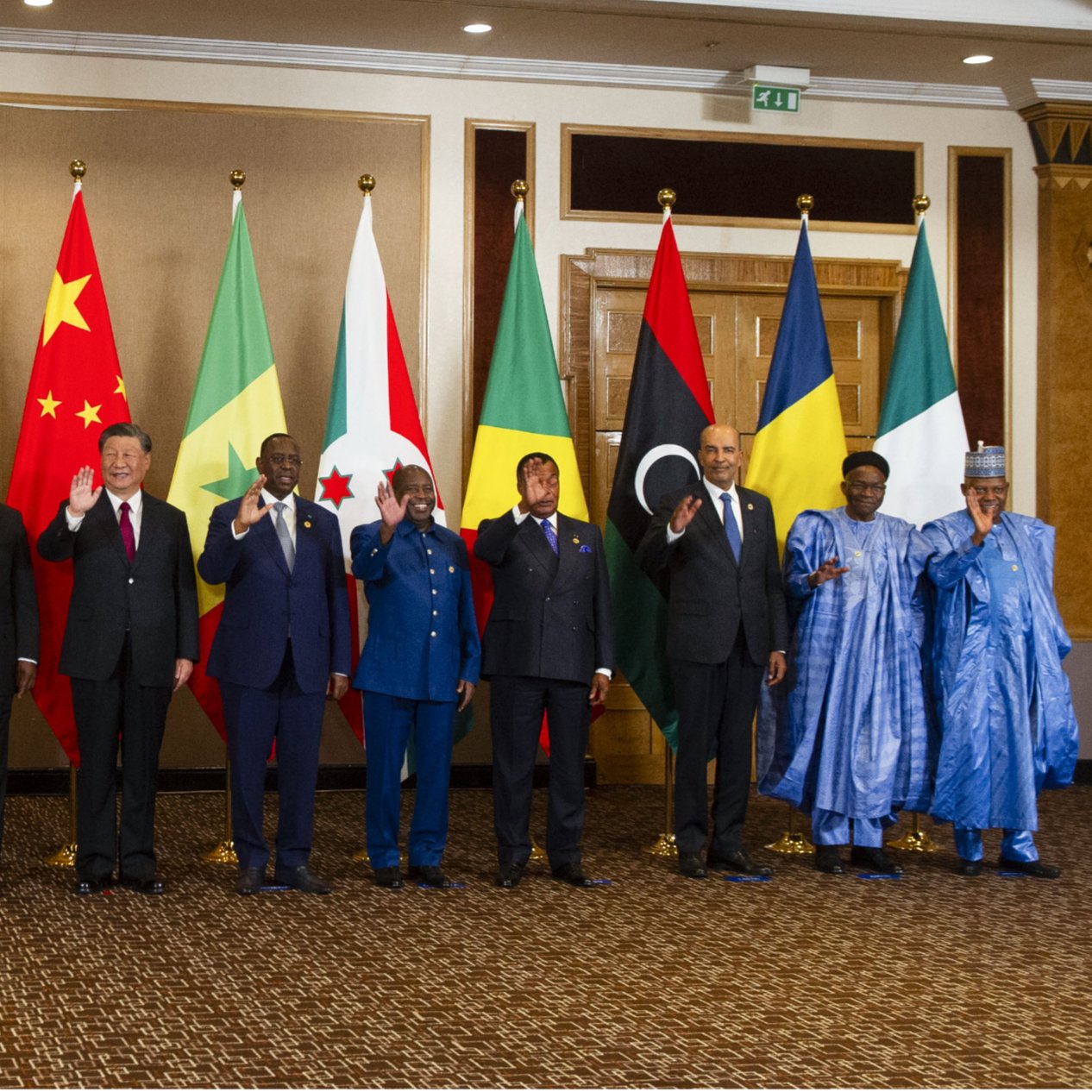 تصویر یک گروه از افراد را نشان می‌دهد که در کنار هم ایستاده‌اند و پرچم‌های مختلف کشورهایشان در پس‌زمینه قرار دارد. به نظر می‌رسد این افراد مقام‌های بلندپایه یا رهبران سیاسی هستند و همگی در حال خوش‌آمدگویی یا دست wave کردن به دوربین هستند. پرچم‌های مختلف شامل پرچم‌های کشورهای آفریقایی و همچنین پرچم‌های چین و سایر کشورها وجود دارد. فضای تصویر رسمی به نظر می‌رسد و افراد لباس‌های رسمی بر تن دارند.