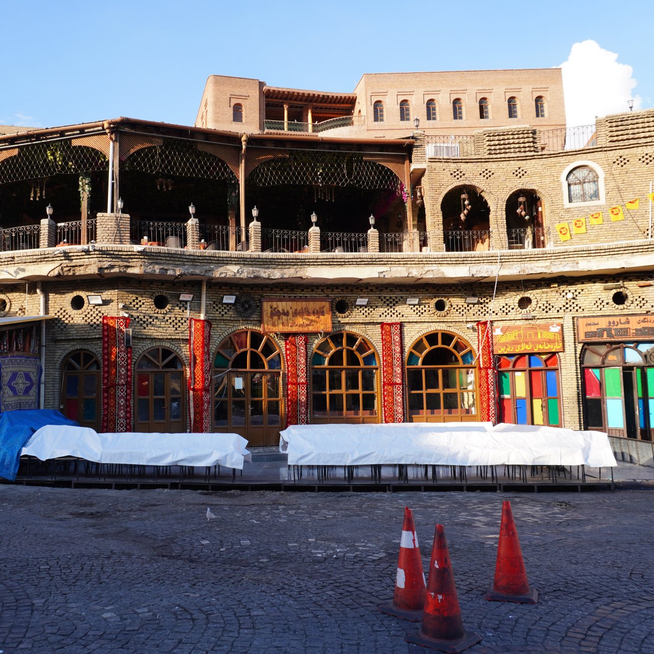 The image depicts a beautifully constructed building featuring intricate architectural details, likely in a historic or culturally significant area. The facade showcases ornate designs, colorful window frames, and a balcony adorned with wooden railings. The ground level appears to have some protective coverings, possibly due to ongoing renovations or maintenance. Surrounding the building, there are orange traffic cones indicating restricted access in that area. The overall ambiance suggests a blend of tradition and vibrant community life.