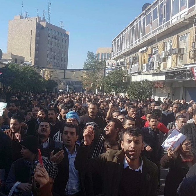 L'image montre une foule dense de personnes rassemblées dans une rue, manifestant visiblement. Les gens tiennent des pancartes et semblent exprimer des revendications. En arrière-plan, on peut voir des bâtiments urbains, ce qui indique que cela se déroule dans une ville. L'atmosphère semble énergique, avec des manifestants qui lèvent les bras et chantent. Les manifestants portent des vêtements variés, et on peut apprécier la diversité des participants.